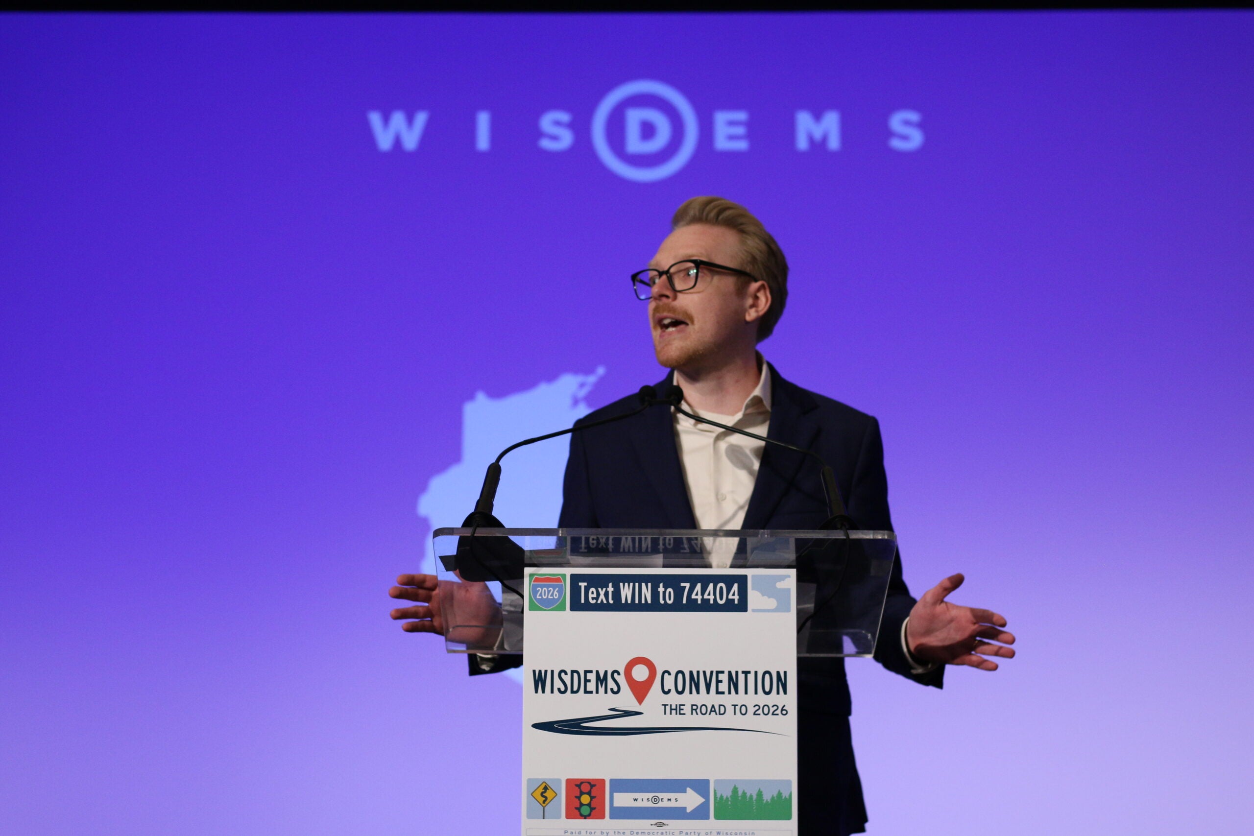 A person speaks at a podium during the WisDems Convention, with a microphone and a projected WisDems logo in the background.
