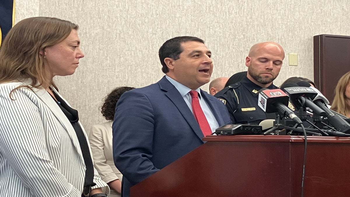 A man in a suit speaks at a podium with microphones, flanked by a woman and a uniformed police officer, during a press conference indoors.