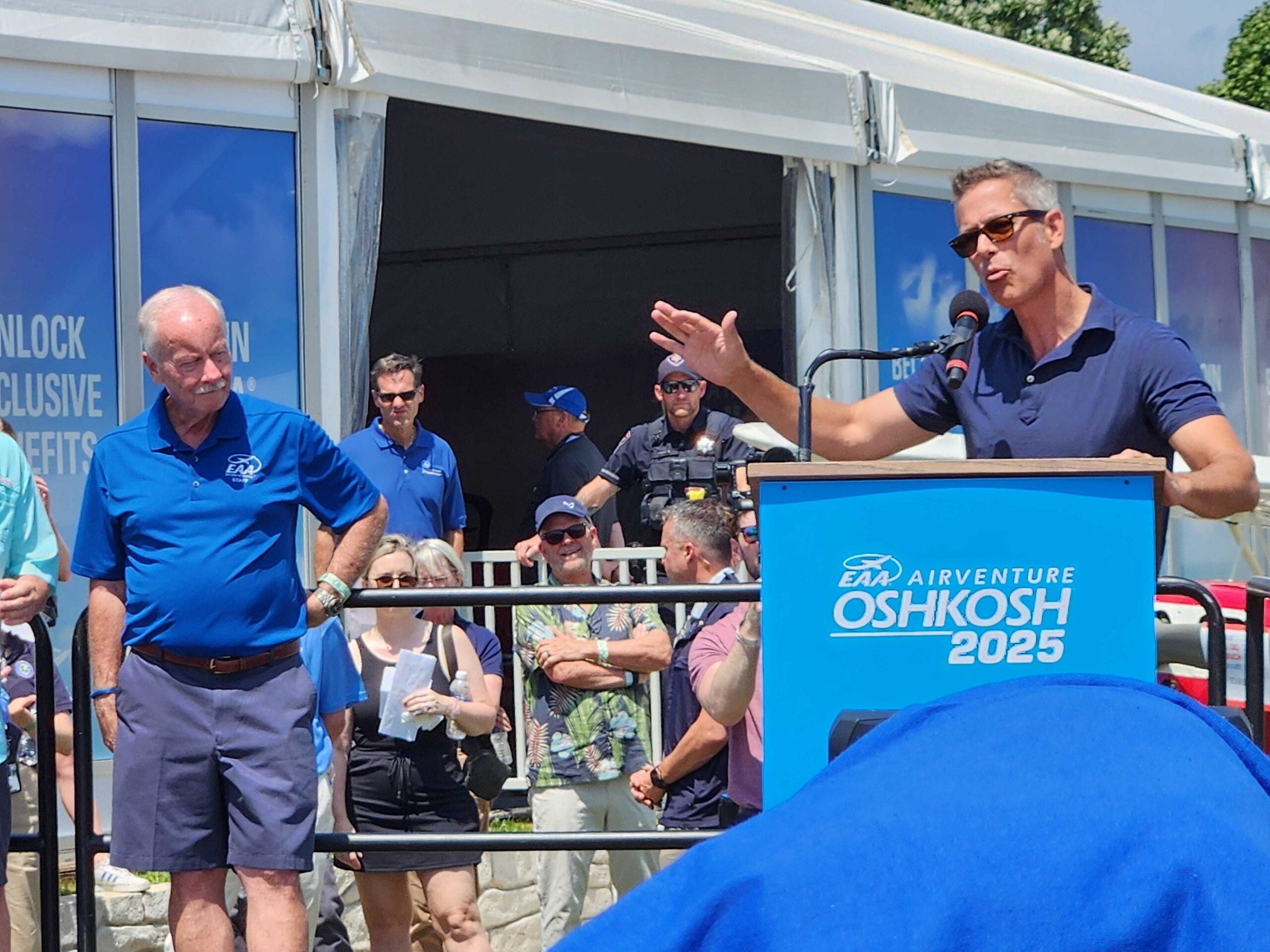 A man speaks at a podium labeled “EAA AirVenture Oshkosh 2025” outdoors, while another man in a blue shirt stands nearby and a group of people watch in the background.