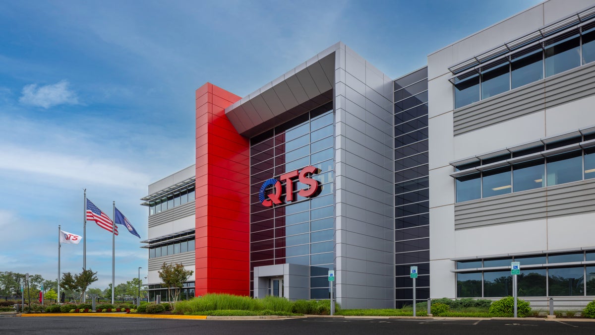 Modern office building with QTS logo on glass facade, featuring red and gray exterior panels, landscaped entrance, and two flags in front.