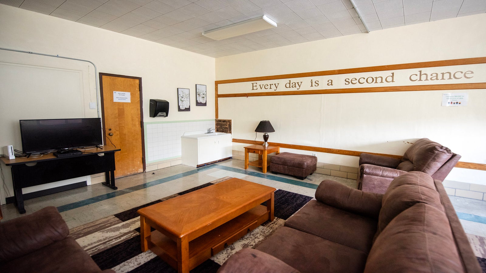 A modest lounge with brown sofas, a wooden coffee table, a TV, and a wall sign that reads, Every day is a second chance.