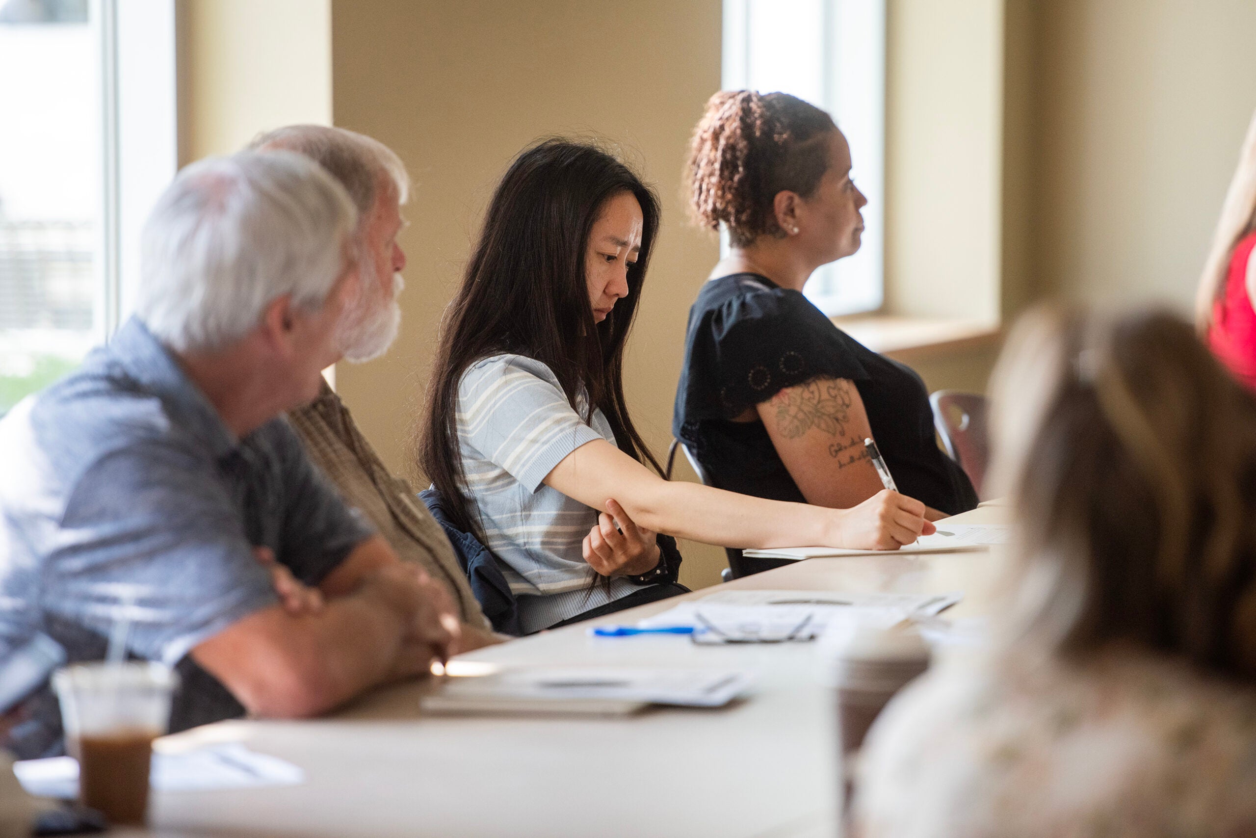 Four adults sit at a table in a well-lit room, listening attentively; one woman in the center is writing in a notebook.
