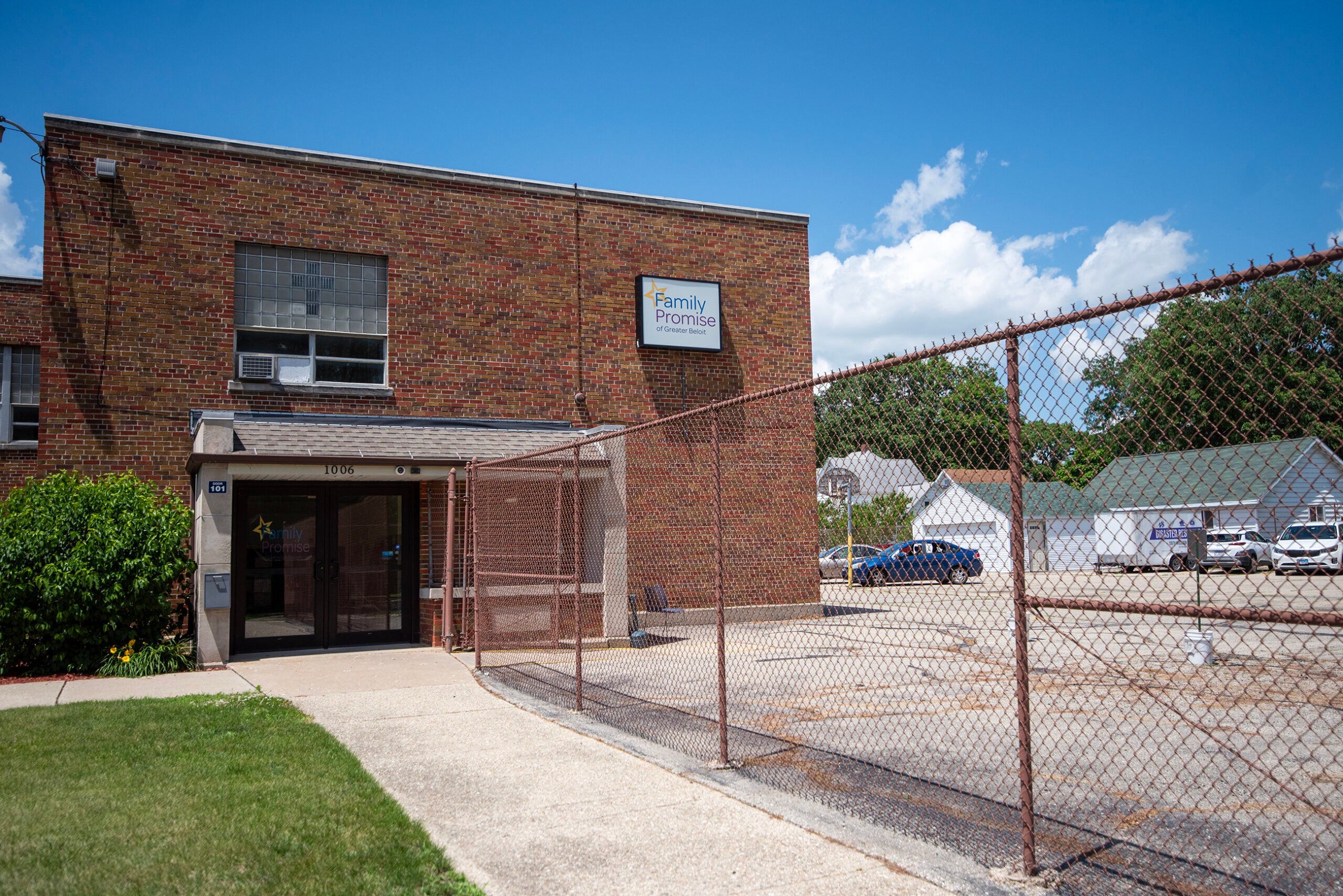 A brick building with a sign reading Family Promise above the entrance, next to a fenced parking lot on a sunny day.