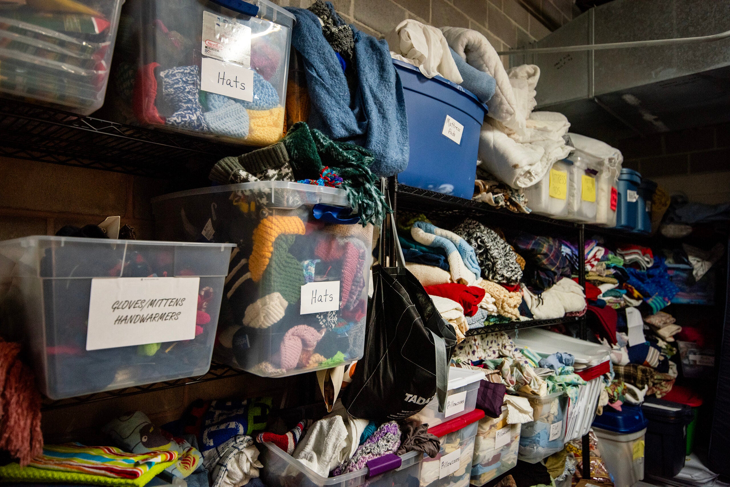 Plastic bins and folded textiles are stored on metal shelves in a utility room. Labels on bins indicate contents such as hats, gloves, mittens, and handwarmers.