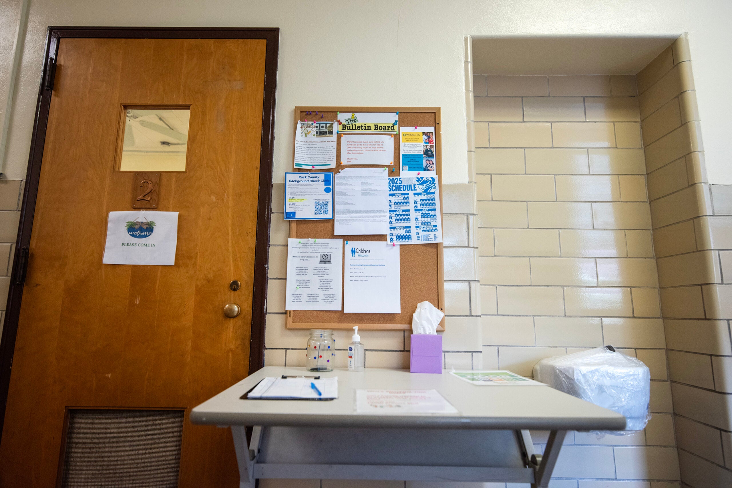 A wooden door and a bulletin board with notices are next to a table holding hand sanitizer, tissues, and papers in a tiled hallway.
