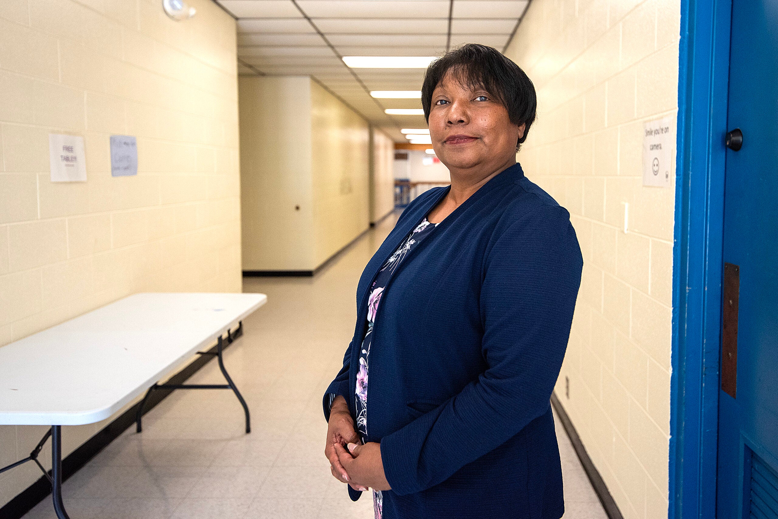 A woman stands in a hallway next to a white folding table, wearing a navy blazer over a floral shirt. The hallway has cream-colored walls and a tiled floor.