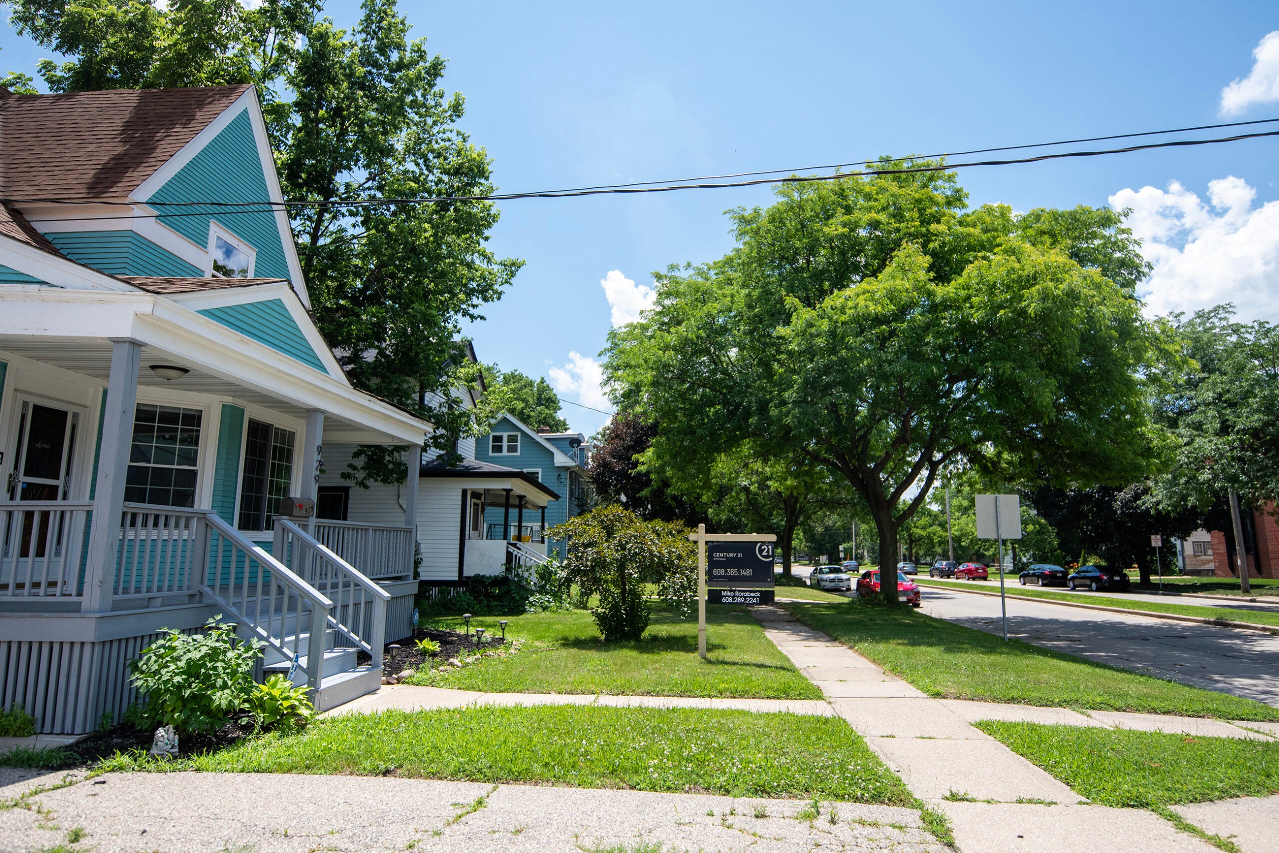 A residential street with houses, trees, a For Sale sign on one lawn, and parked cars under a clear blue sky.