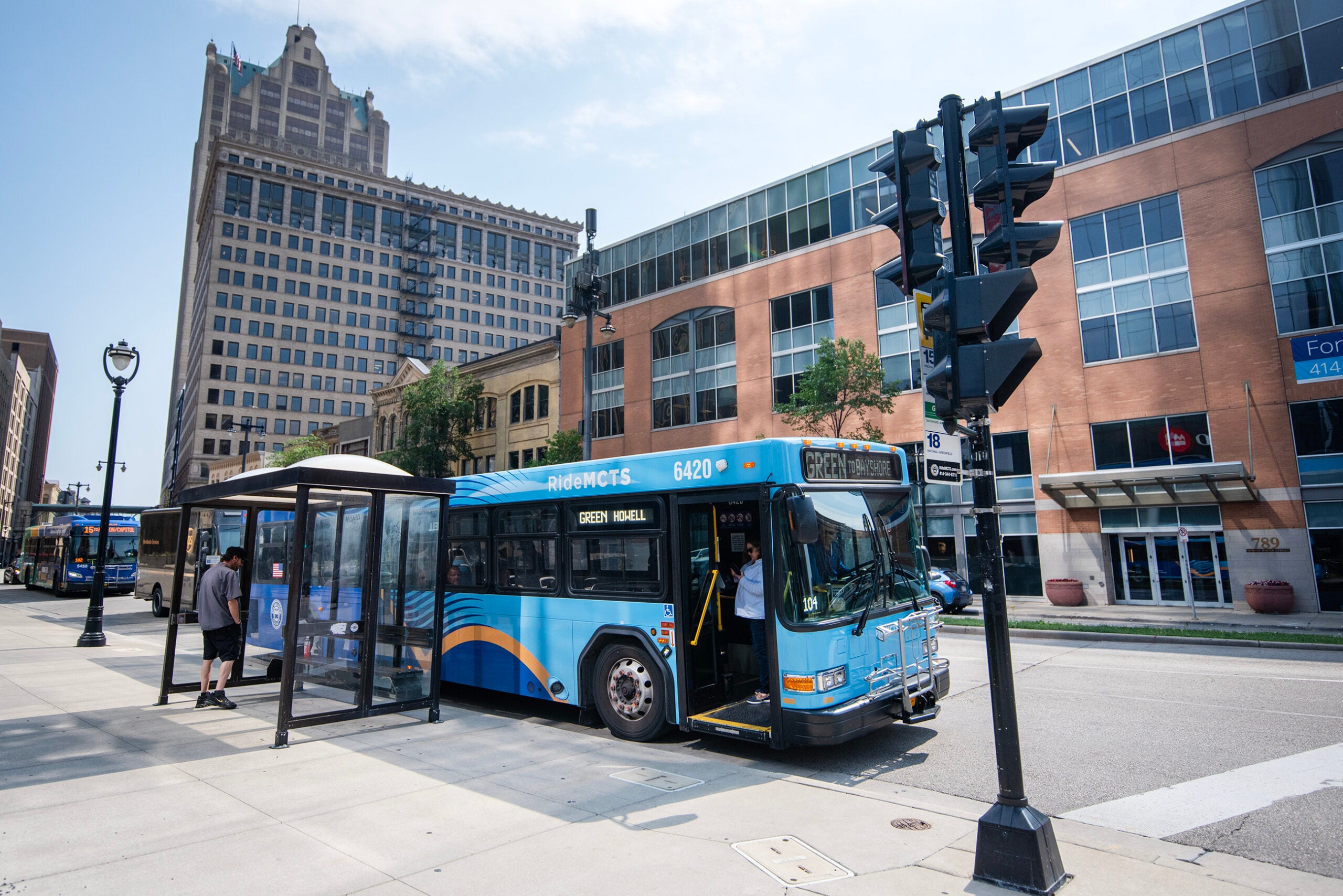A city bus marked Green MEGA stops at a bus shelter on an urban street, with people boarding and buildings in the background.
