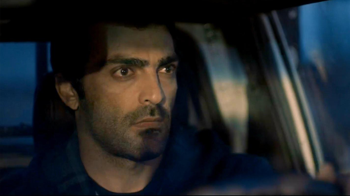 A man with a serious expression sits behind the wheel of a vehicle, focused on the road ahead in dim lighting.