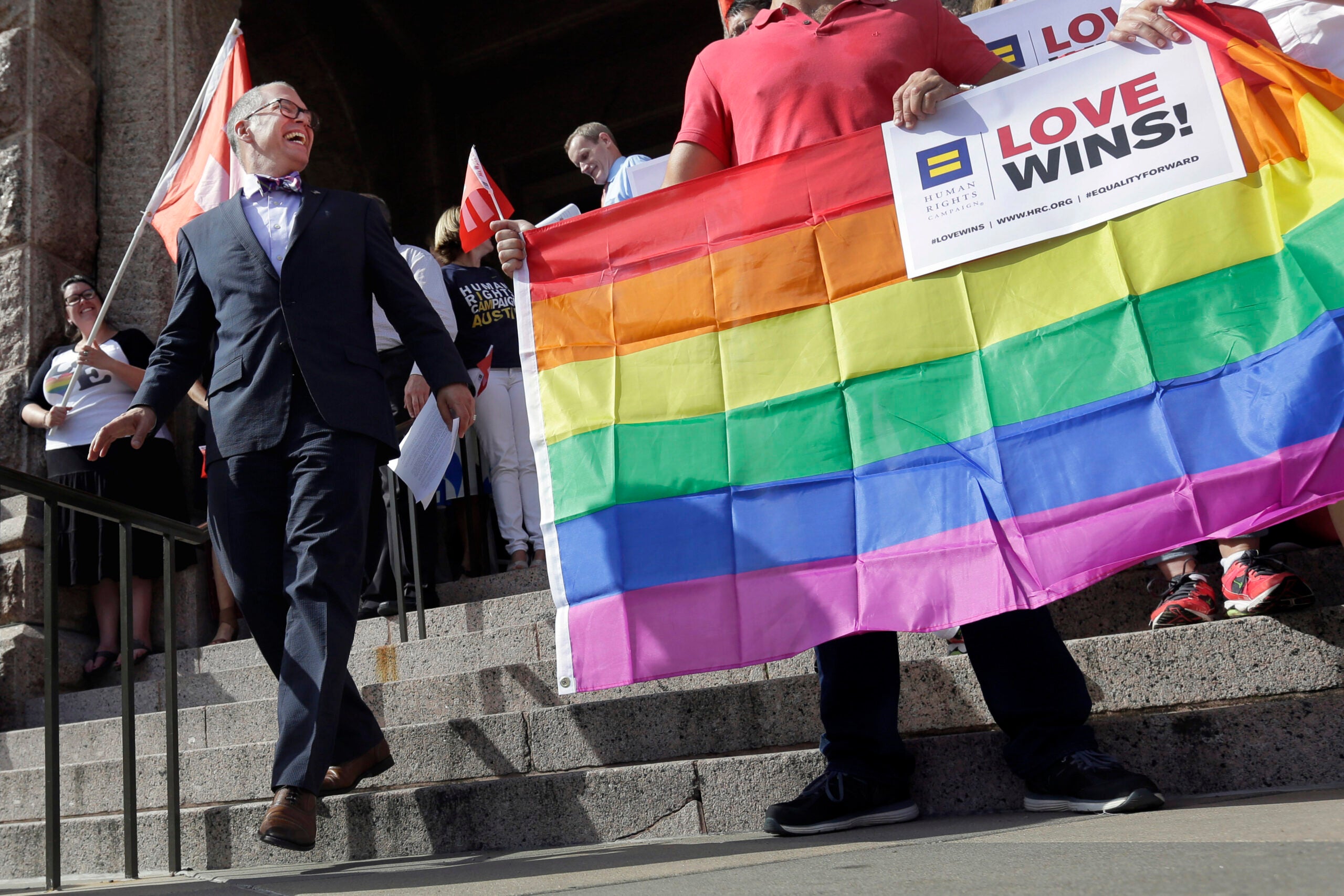 People stand on building steps holding a rainbow pride flag and signs reading Love Wins! during a gathering or rally.