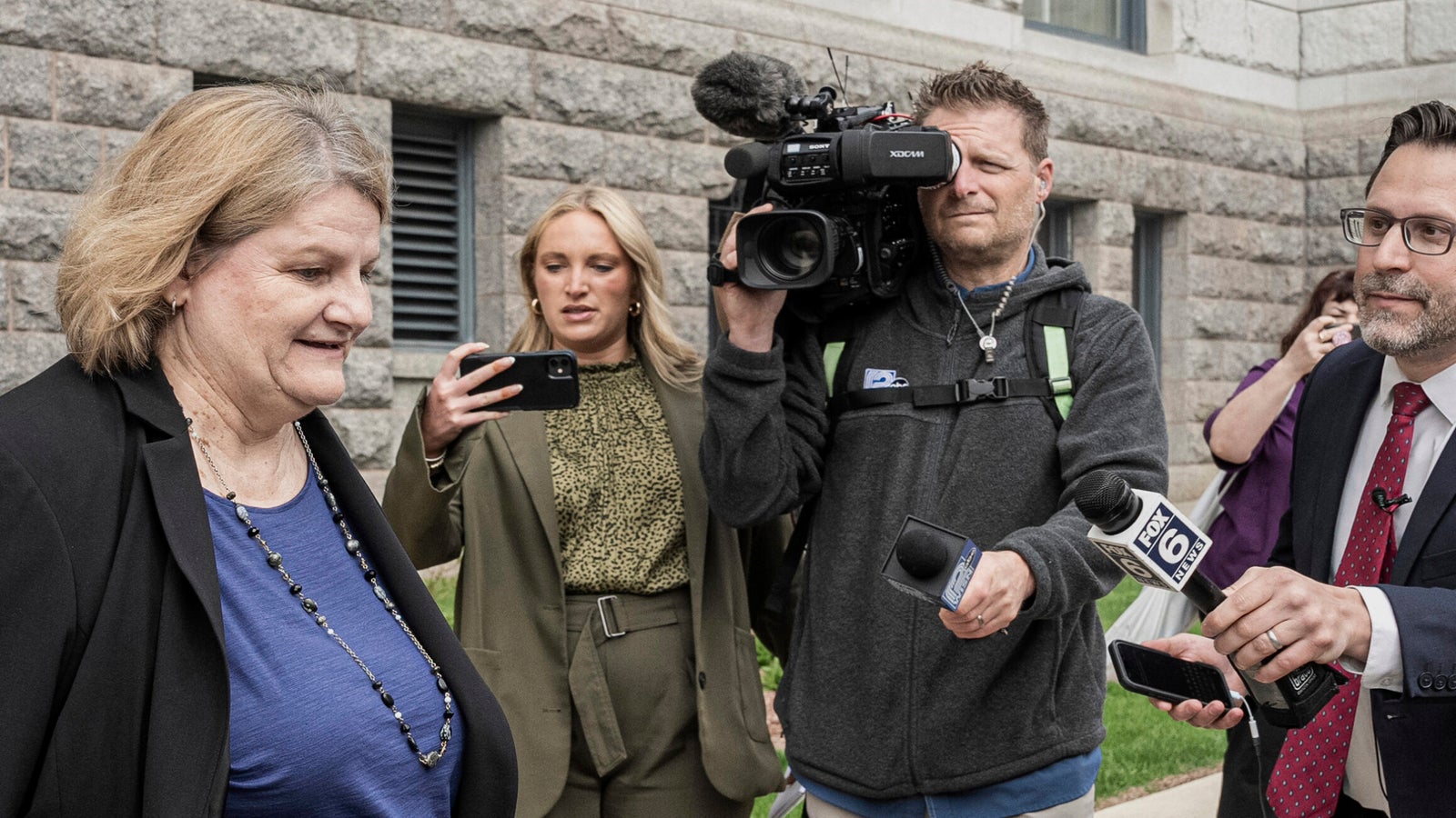 A woman walks outside a stone building while being approached by reporters with microphones and a camera operator filming the scene.