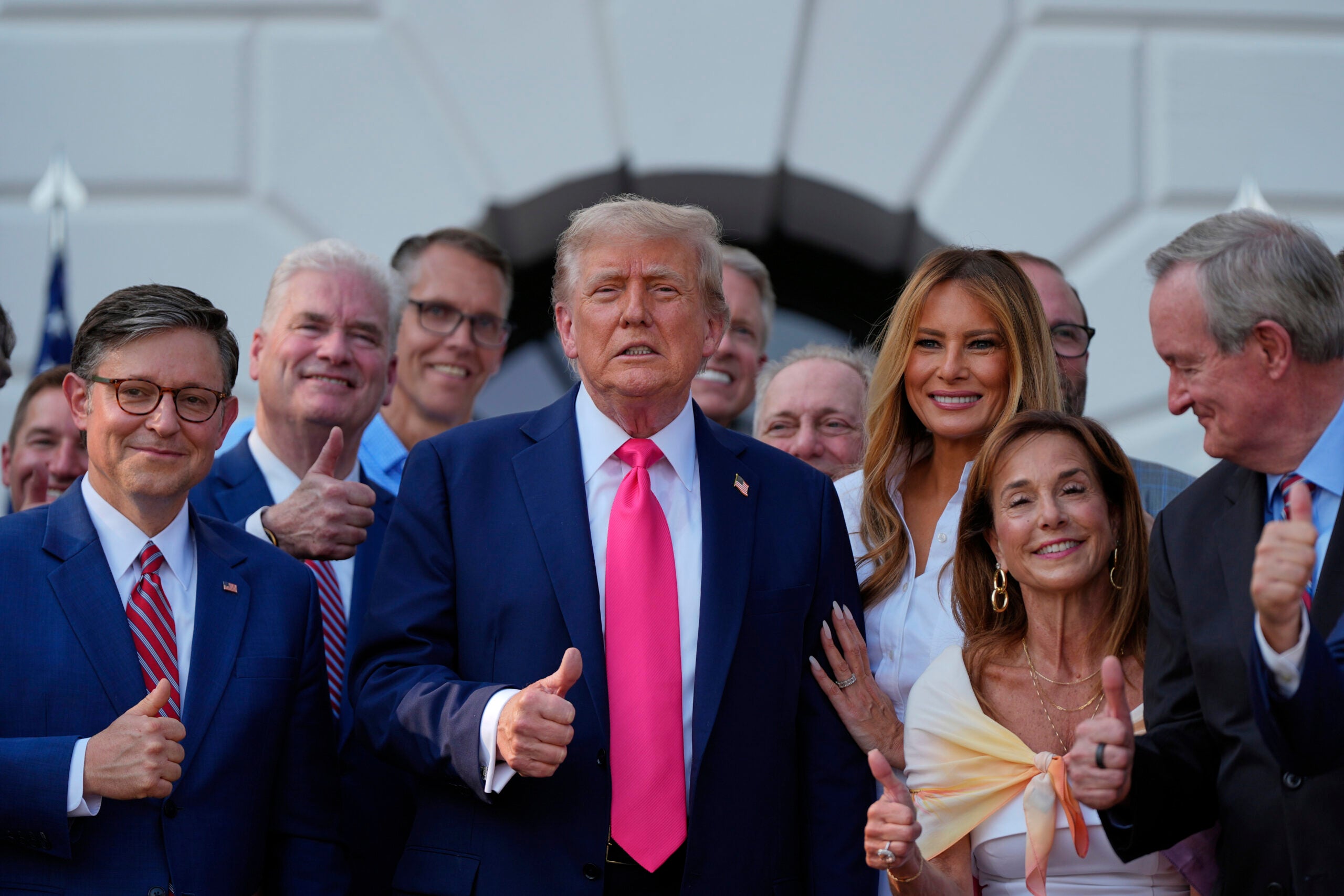 A group of people, including Donald Trump in a suit and pink tie, stand outside a white building, smiling and giving thumbs up.