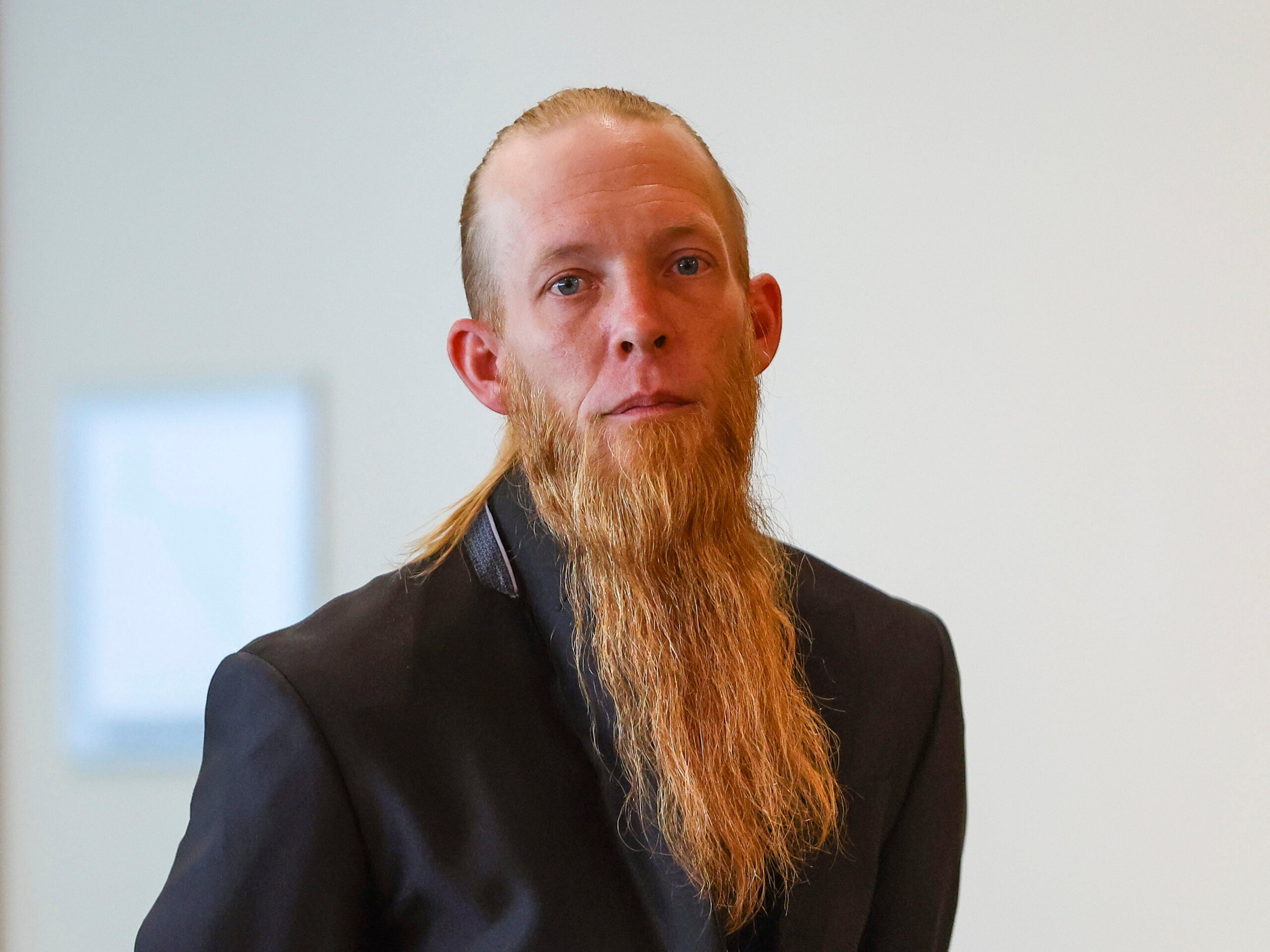 A man with a long reddish-blonde beard and slicked-back hair is wearing a black suit jacket, standing against a plain light background.
