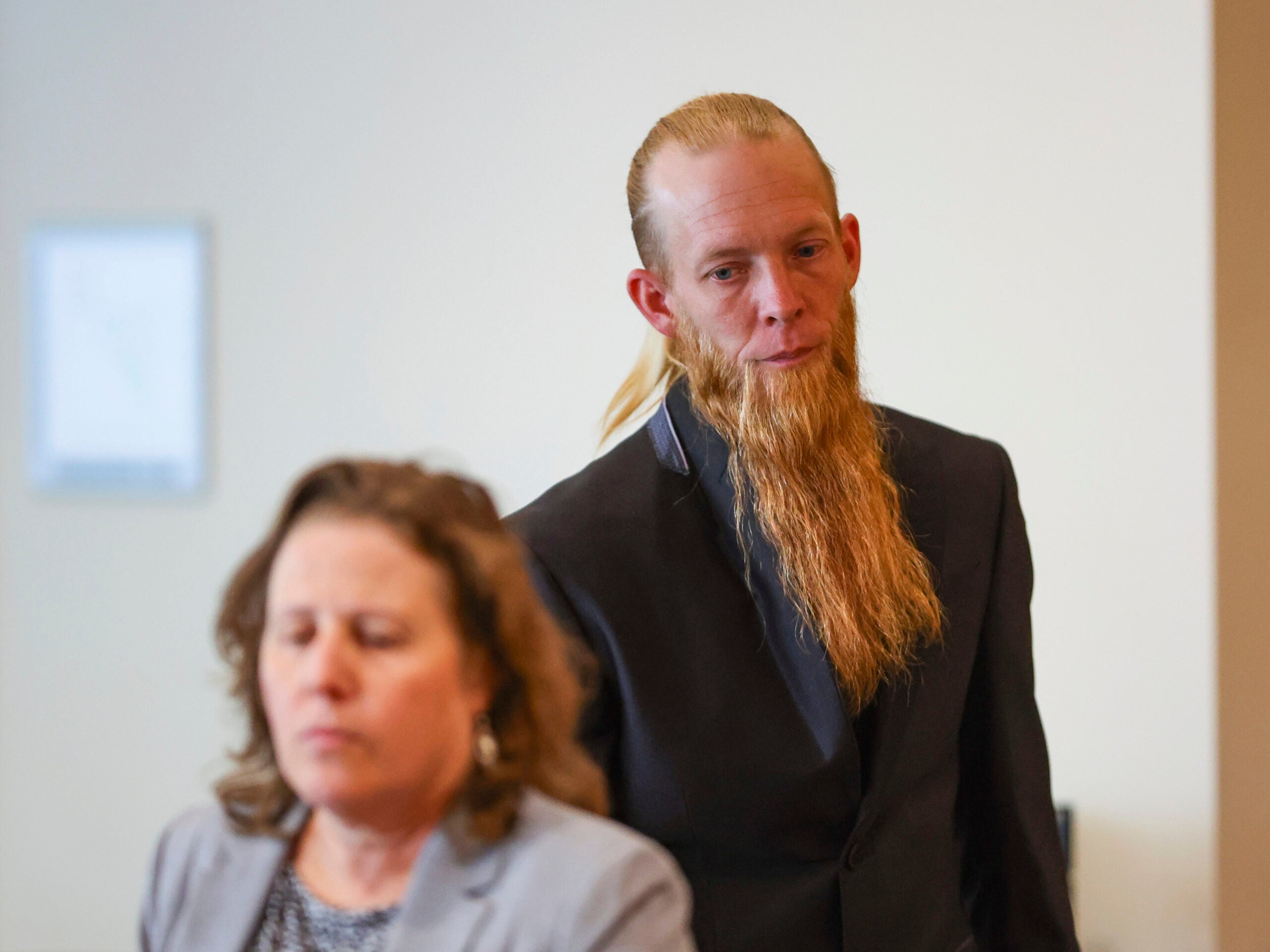 A man with a long beard and a suit stands behind a seated woman in a gray jacket, both indoors in a neutral setting.