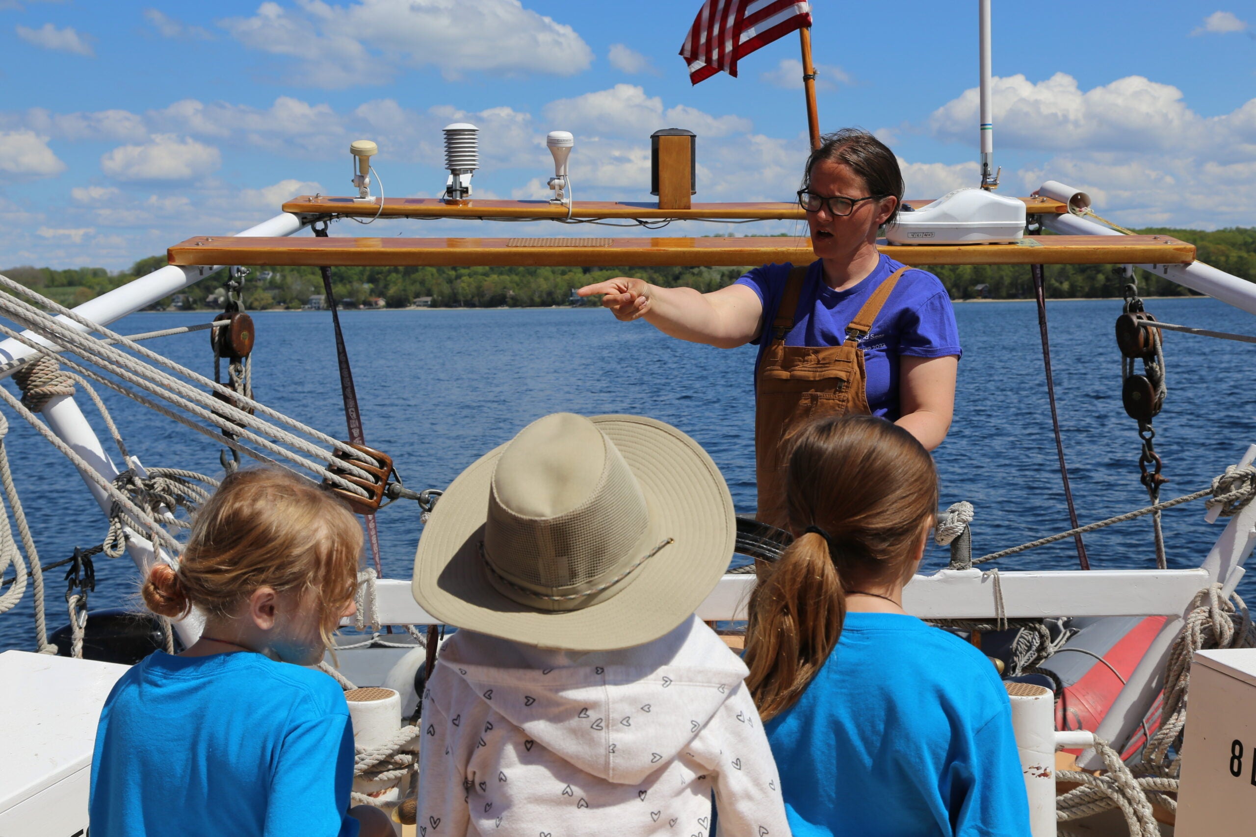 An adult explains something to three children on a boat, with water and trees in the background and an American flag visible above.