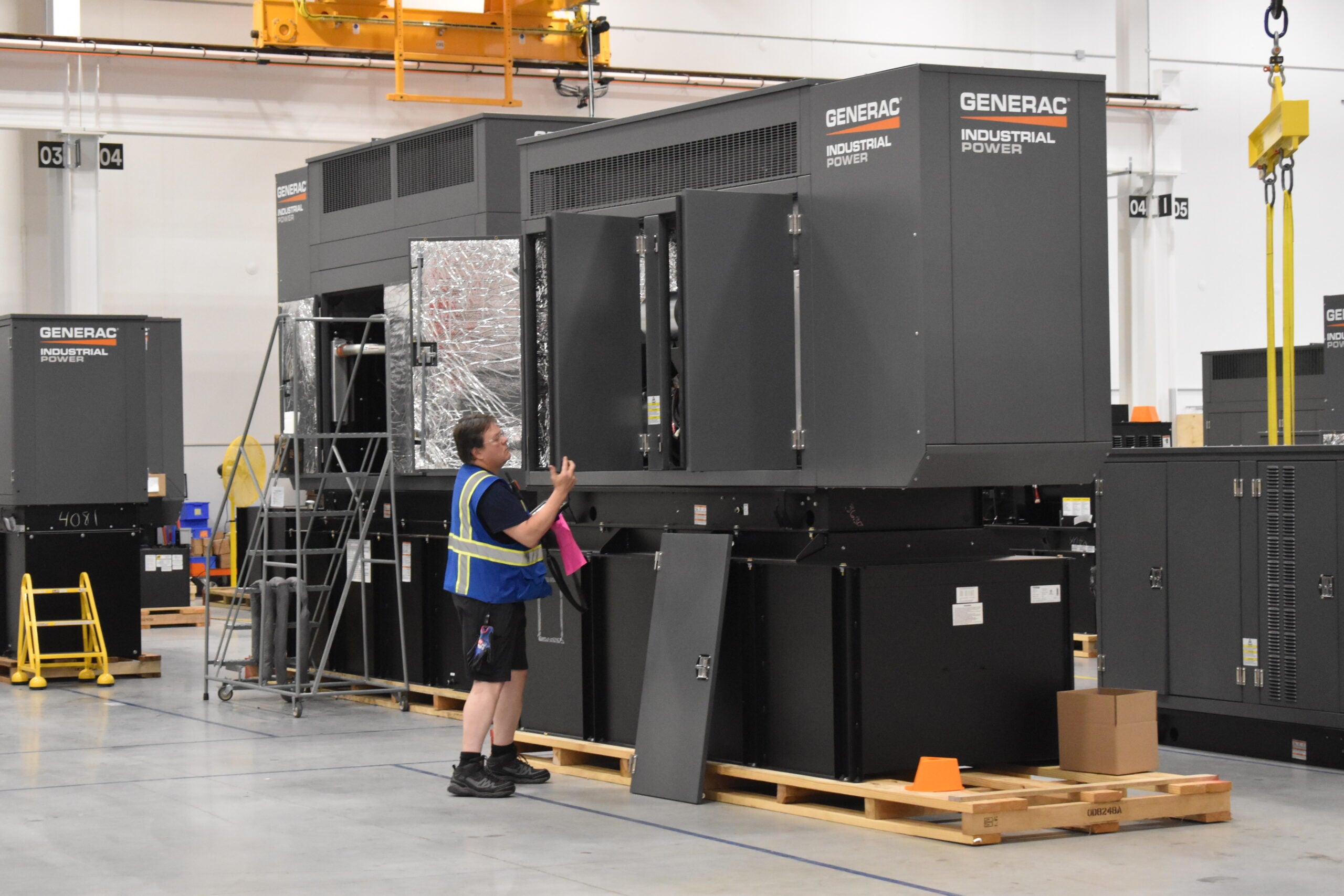 A worker in a safety vest inspects large Generac industrial power generators inside a warehouse.