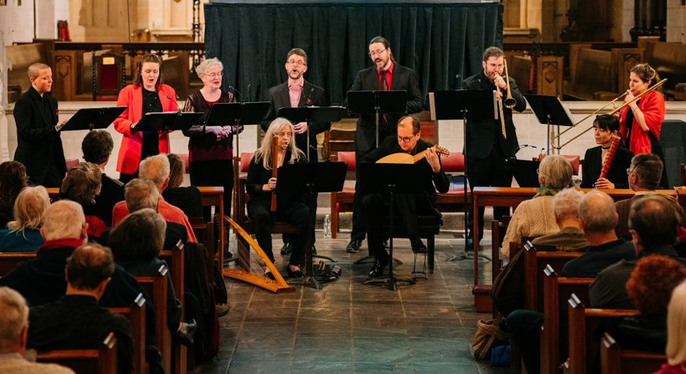 A group of musicians and singers perform on a stage in a church, facing an audience seated in pews.