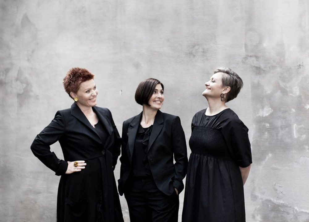 Three women in black outfits stand together against a plain, light gray wall. Two are looking at the woman in the center, who is smiling.