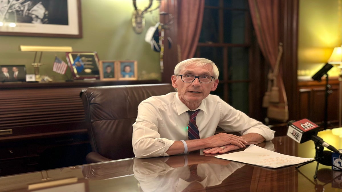 An older man with glasses and a tie sits at a desk with papers in front of him, speaking into microphones in an office with wood paneling and framed photos.