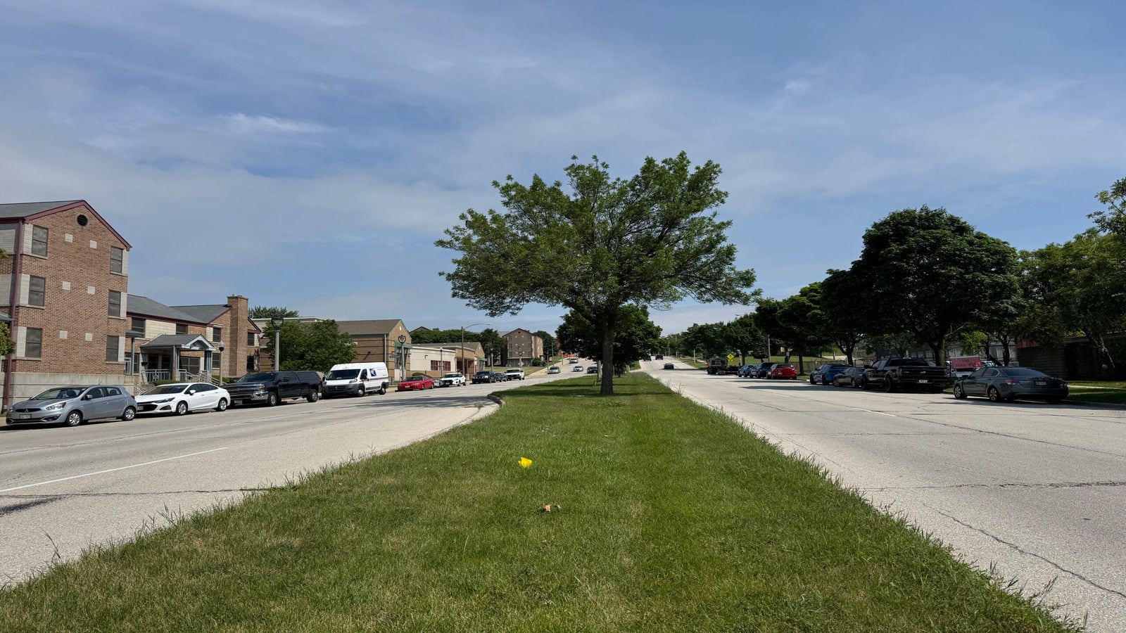 A wide street divided by a grassy median with a tree, flanked by parked cars and residential buildings under a partly cloudy sky.