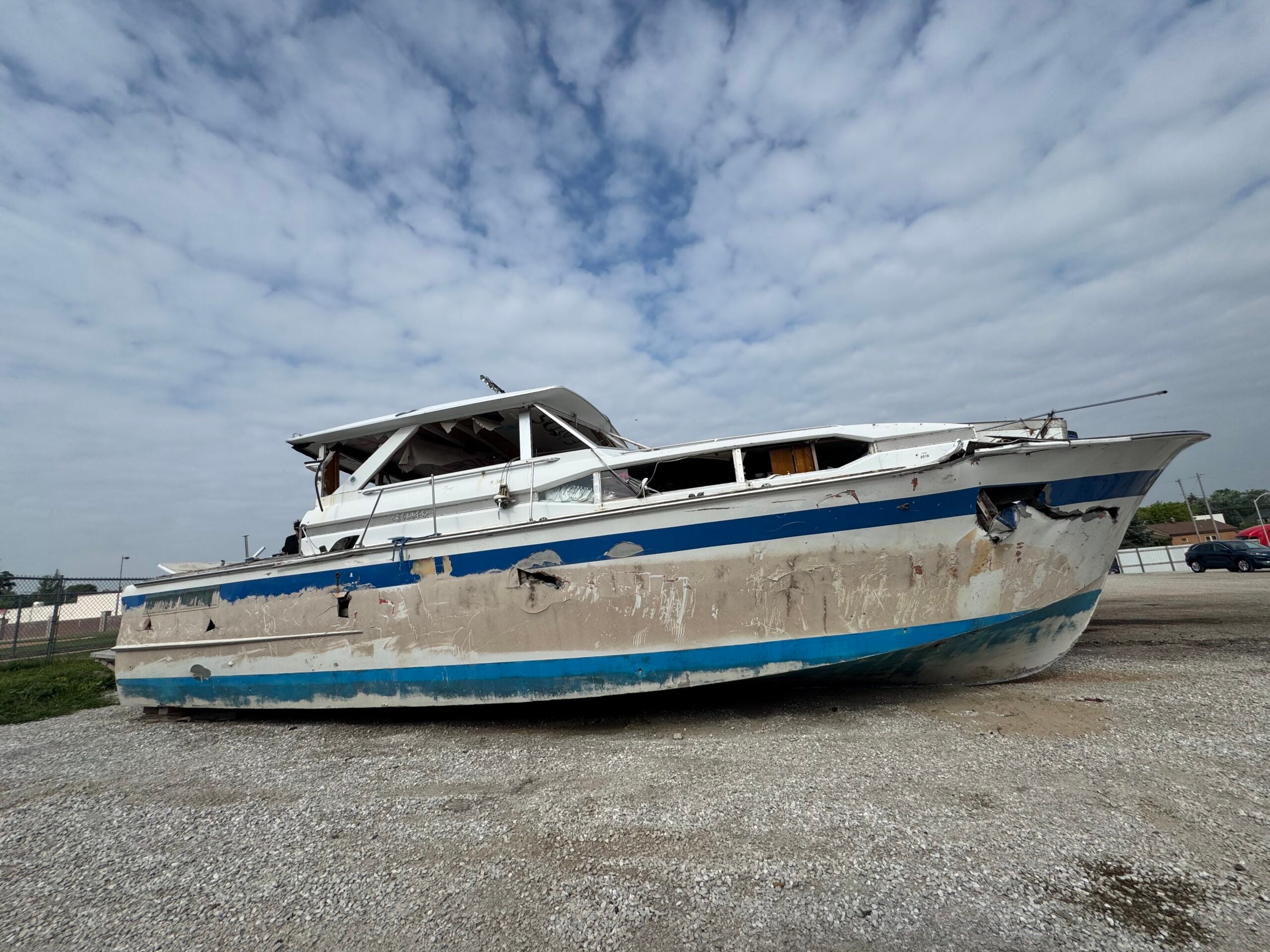 A large, damaged white and blue boat sits on gravel under a cloudy sky, with visible dents and peeling paint along the hull.