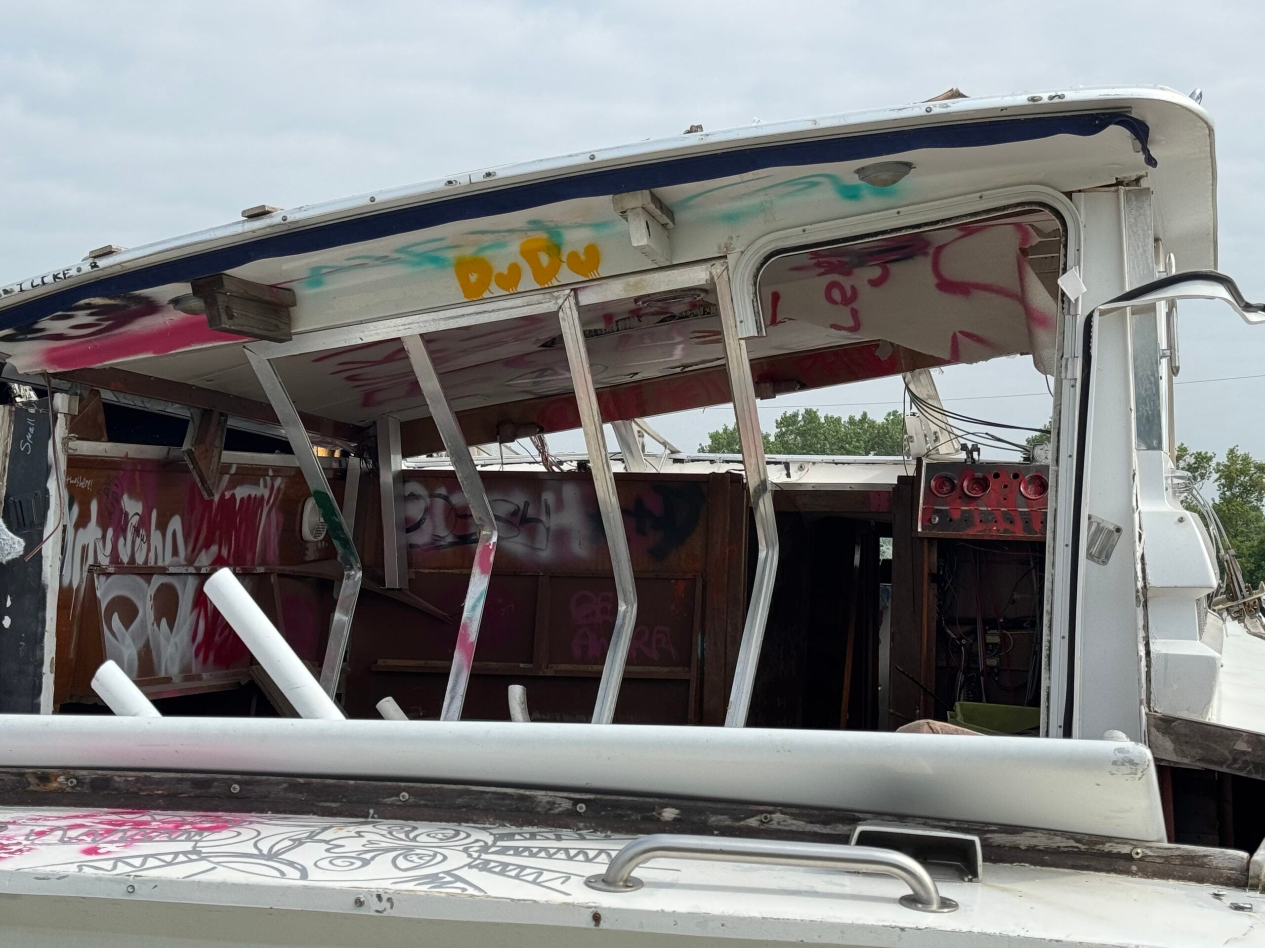 A partially destroyed and heavily graffitied boat with broken windows and exposed interior, set outdoors under a cloudy sky.