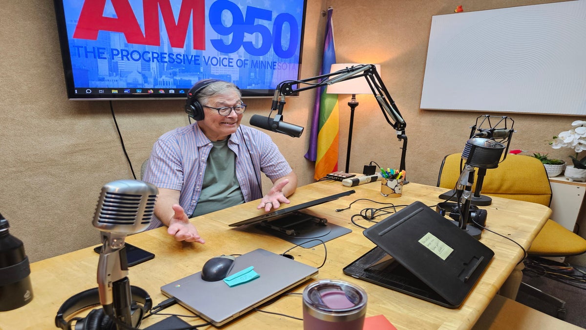 A person wearing headphones and speaking into a microphone sits in a radio studio with a screen showing “AM950” and a rainbow flag in the background.