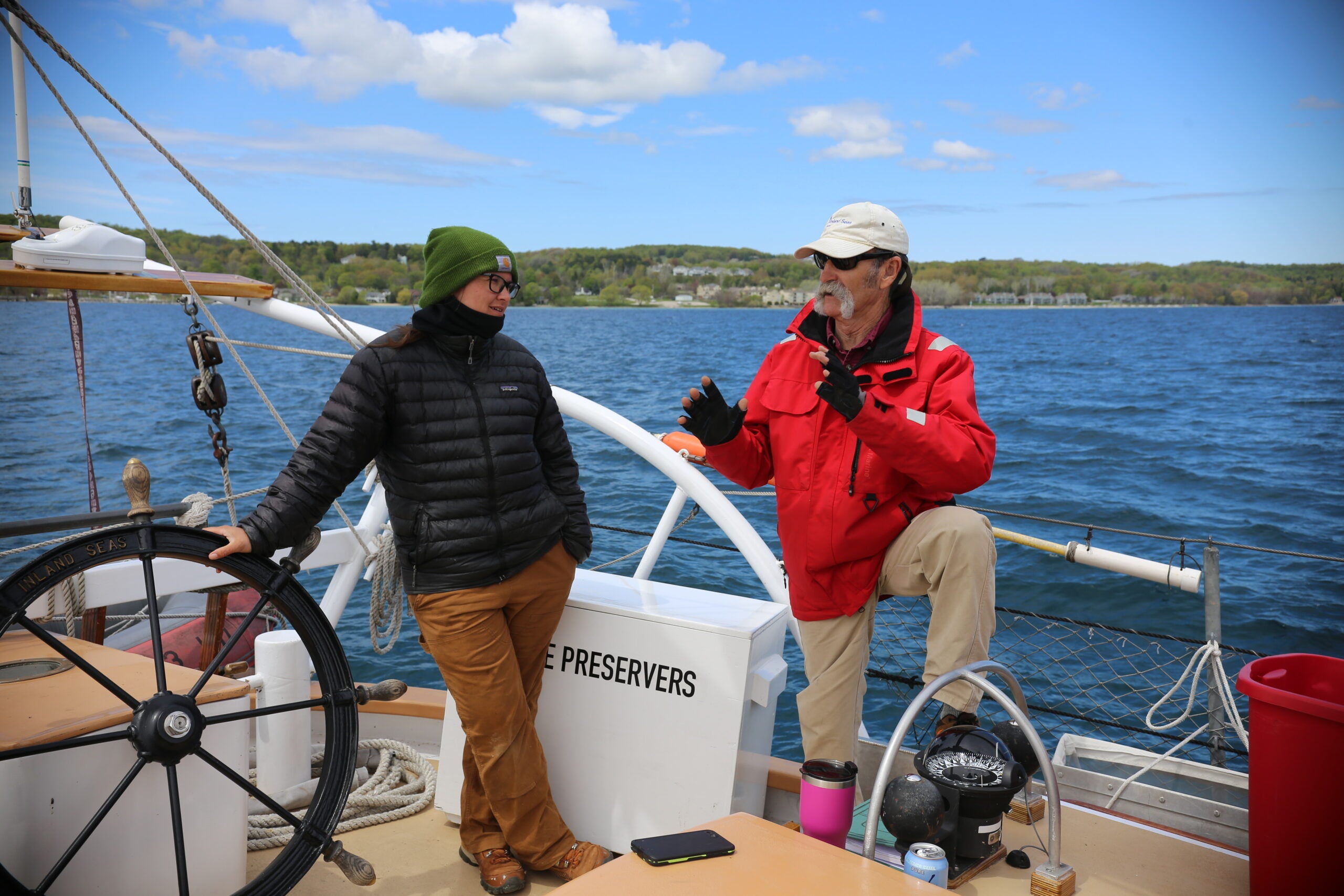 Two people in jackets talk on the deck of a sailboat near a lake or river, with trees and buildings visible in the background under a partly cloudy sky.