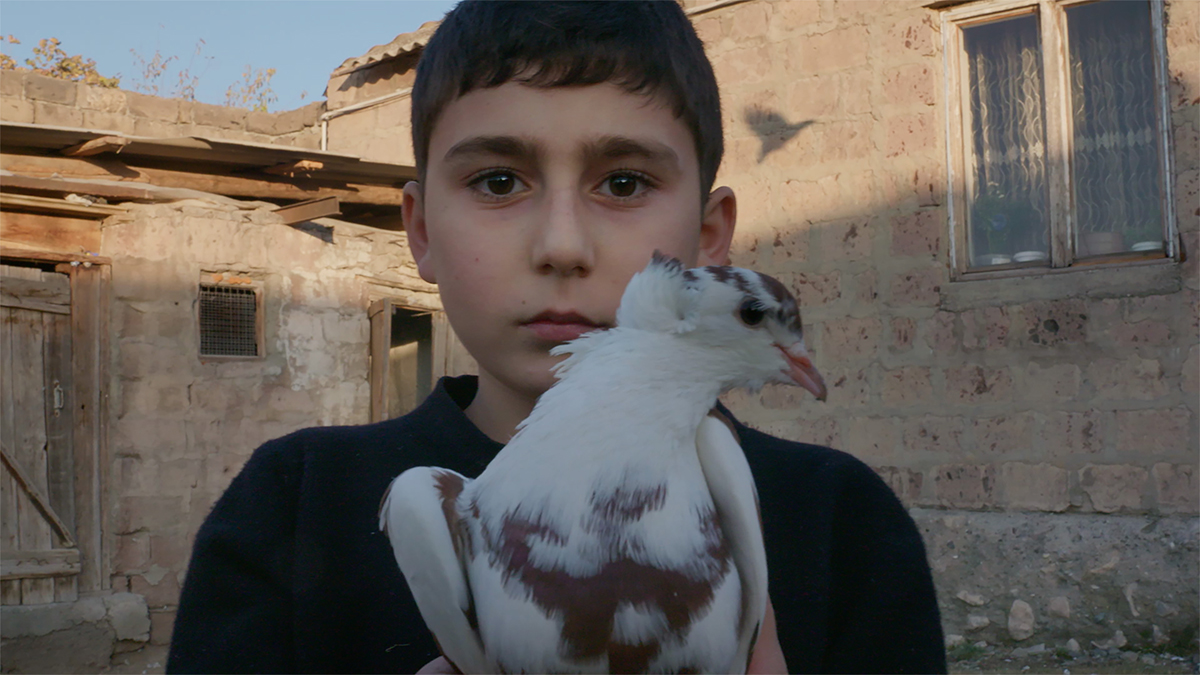 A boy stands outside a stone building, holding a white pigeon with brown markings in his hands and looking directly at the camera.