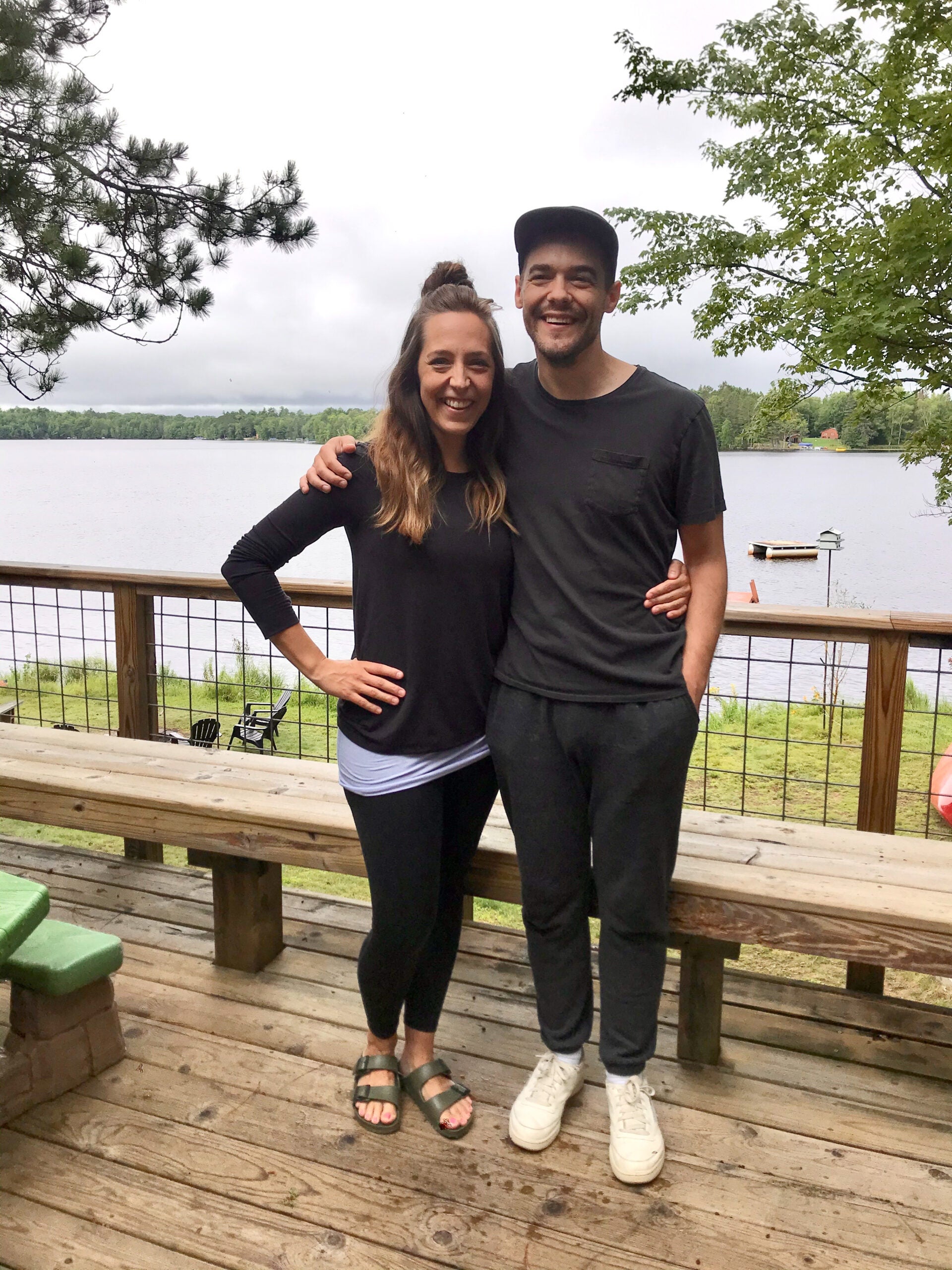 Two people stand side by side on a wooden deck by a lake, smiling at the camera. Trees and cloudy sky are visible in the background.