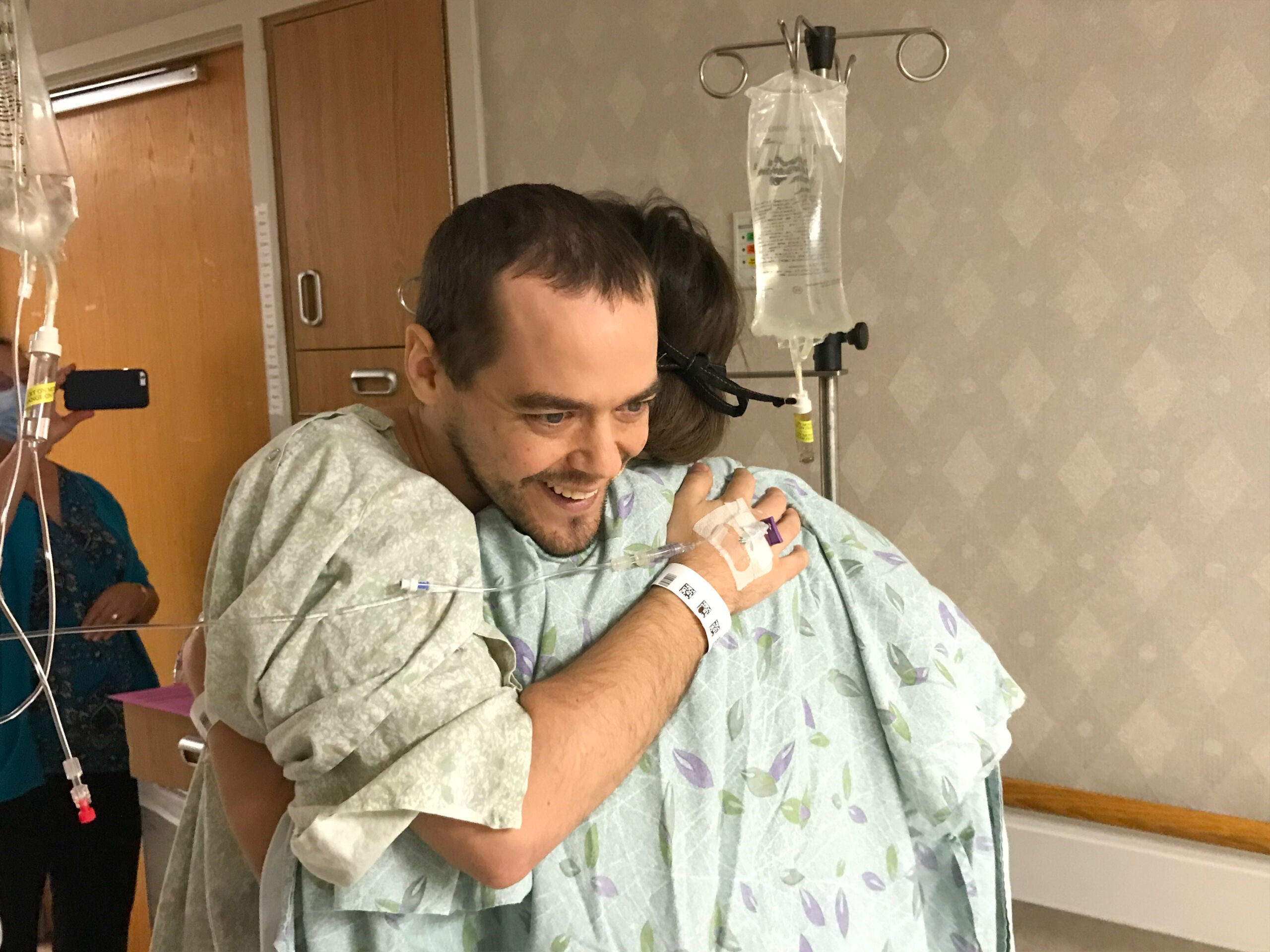 Two people in hospital gowns embrace in a hospital room, with IV stands visible in the background.