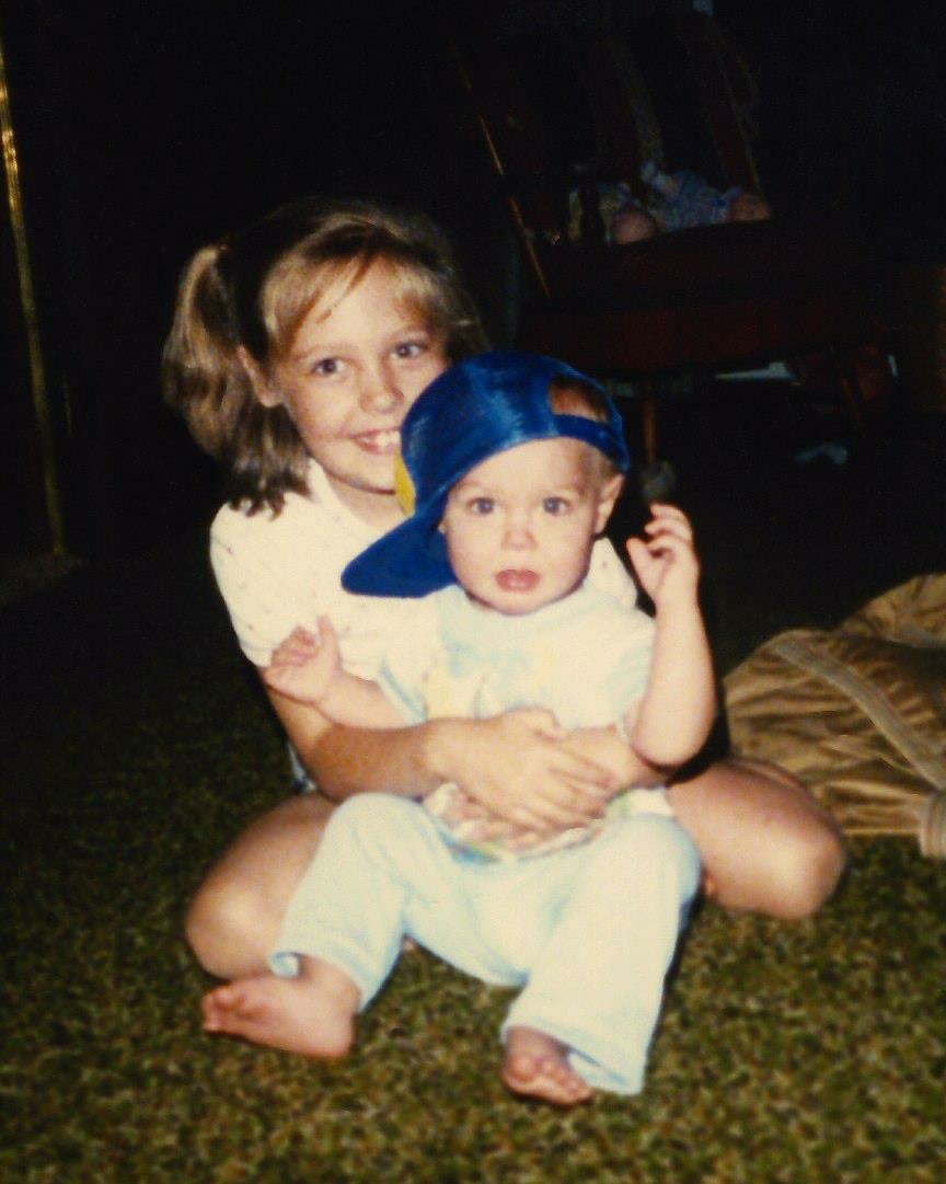 A young girl sits on the floor holding a baby wearing a blue baseball cap sideways. Both children are barefoot and looking at the camera.