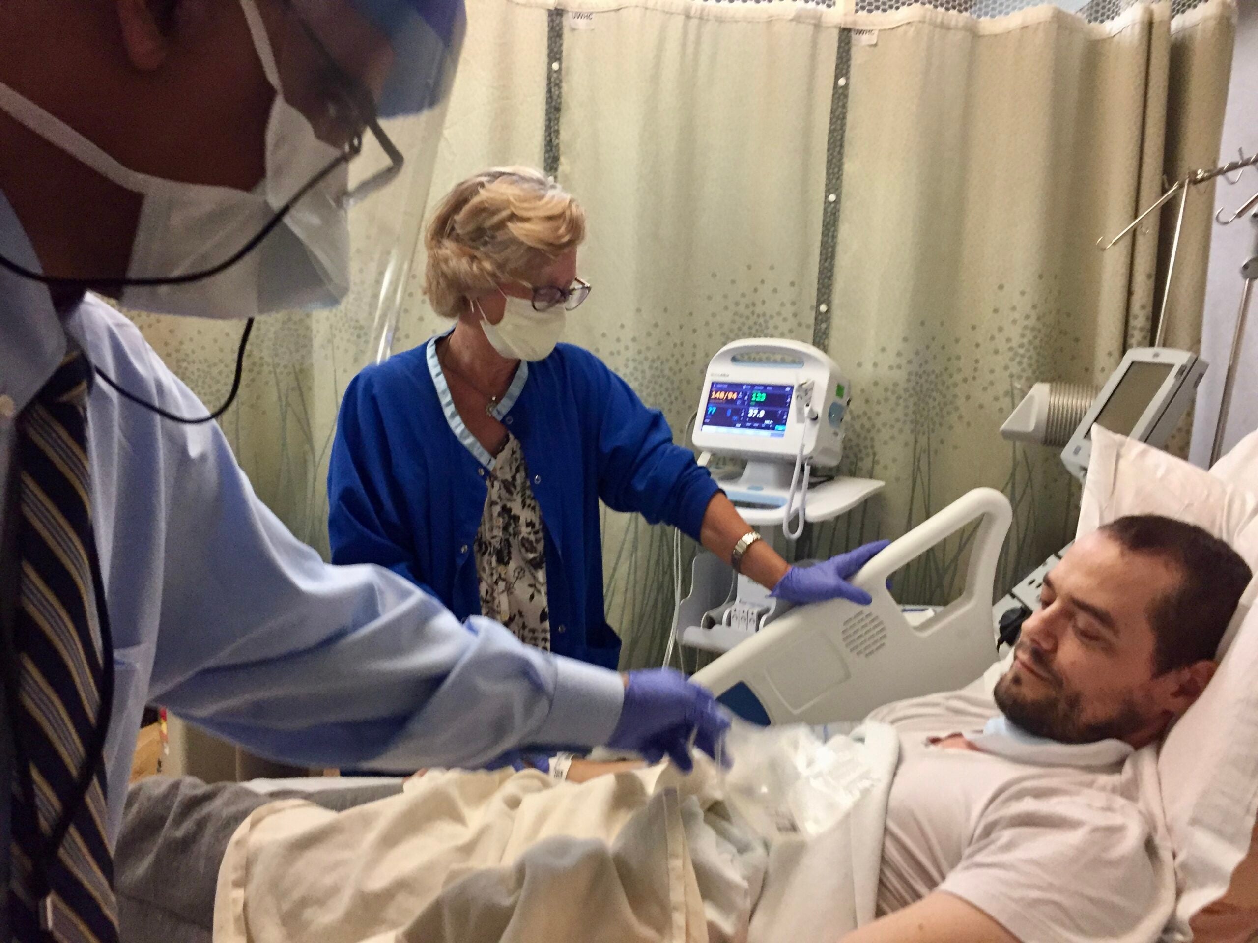 A man lies in a hospital bed while two healthcare workers in protective masks and gloves attend to him in a medical setting.