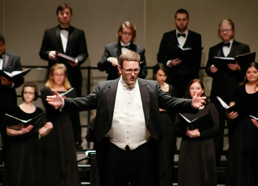 A choir conductor stands at the front with arms outstretched, leading a group of singers dressed in formal black attire on stage.