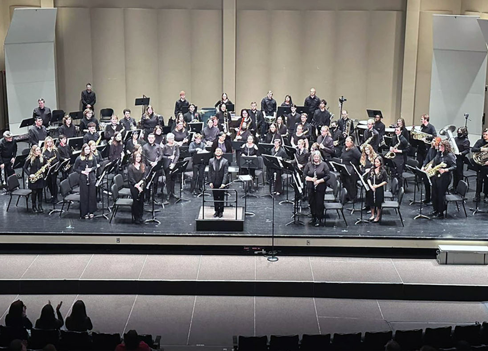 A concert band sits onstage in a large auditorium, with a conductor standing at the podium and audience members seated in front.