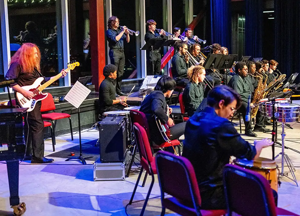 A jazz band performs on stage with musicians playing brass, woodwind, guitar, bass, and percussion instruments, all seated and dressed in black.