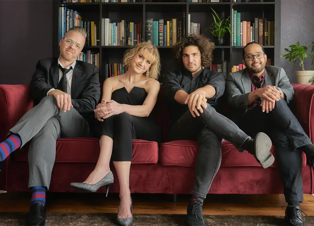Four people sit on a red couch in front of a bookshelf, dressed in semi-formal attire, all looking at the camera with relaxed expressions.