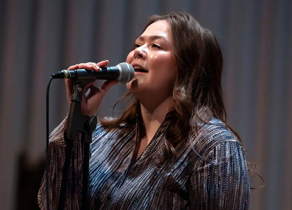 A woman with long brown hair sings into a microphone on stage, wearing a patterned blouse and standing in front of a blurred background.
