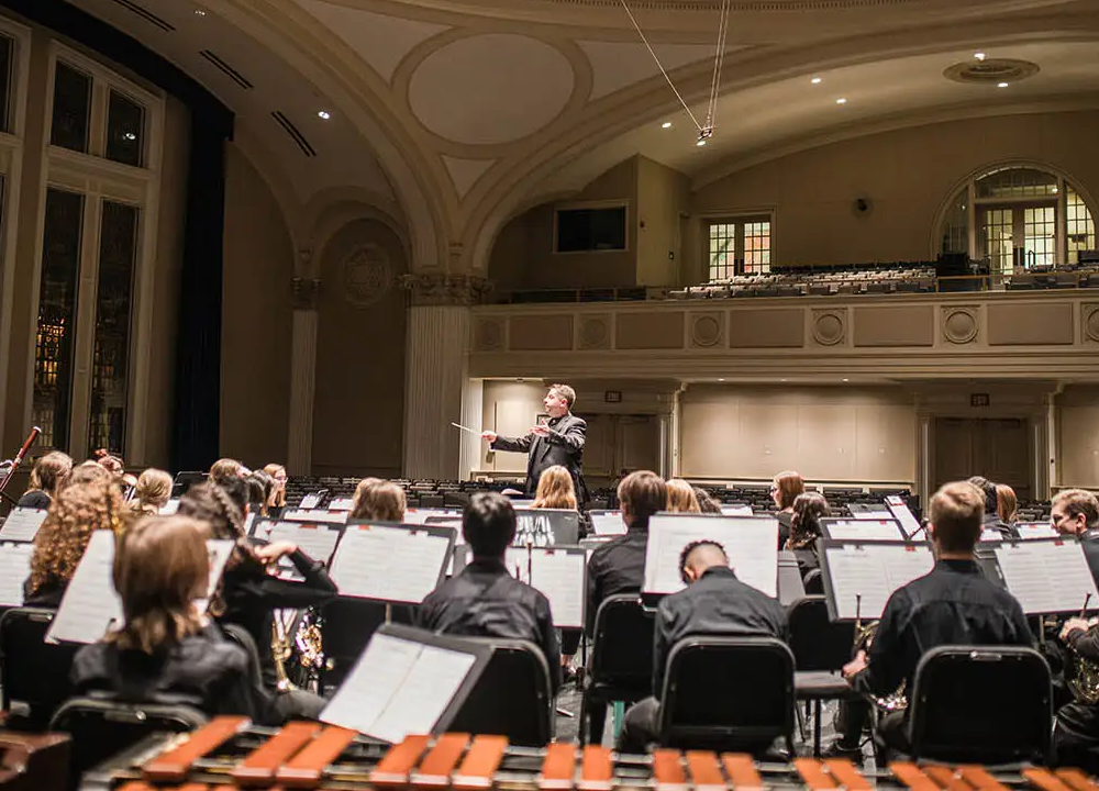 A conductor leads an orchestra of musicians seated with music stands in a large concert hall.