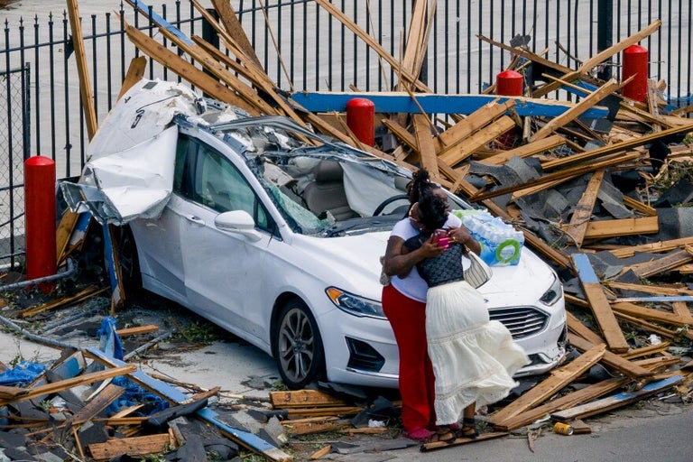 Two people embrace in front of a destroyed car surrounded by debris and broken wooden planks near a metal fence.