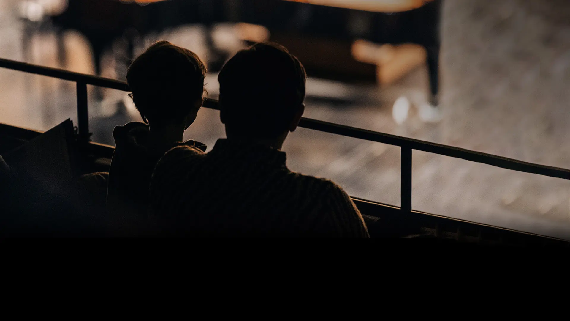 Two people sit side by side in a darkened theater balcony, silhouetted against the stage as they watch a performance below.