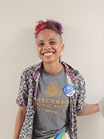 A person with short, dyed hair smiles at the camera. They are wearing a patterned shirt over a Wisconsin Public Radio T-shirt and a round button.