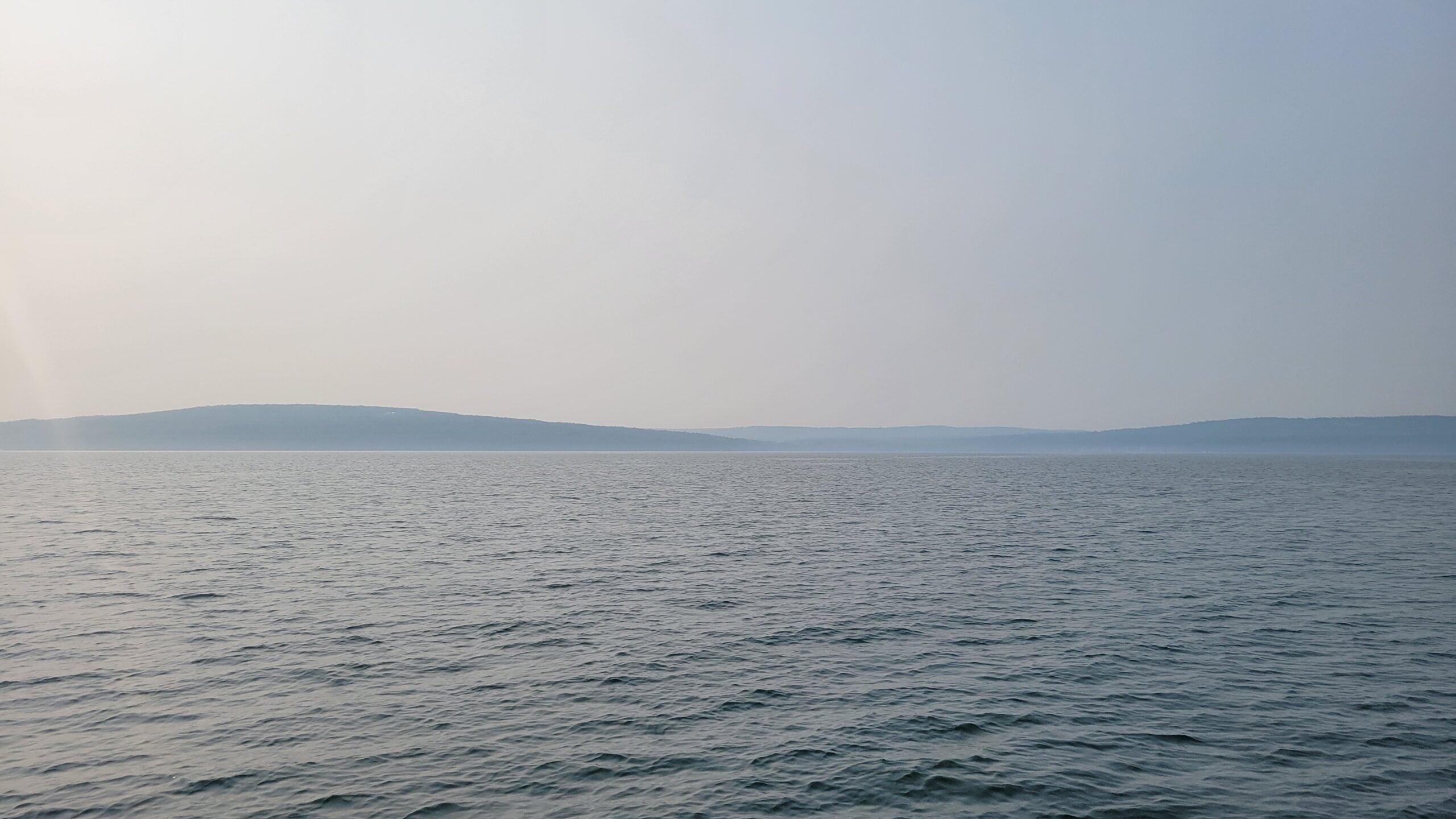 A calm, expansive body of water under a hazy sky with distant, low hills on the horizon.