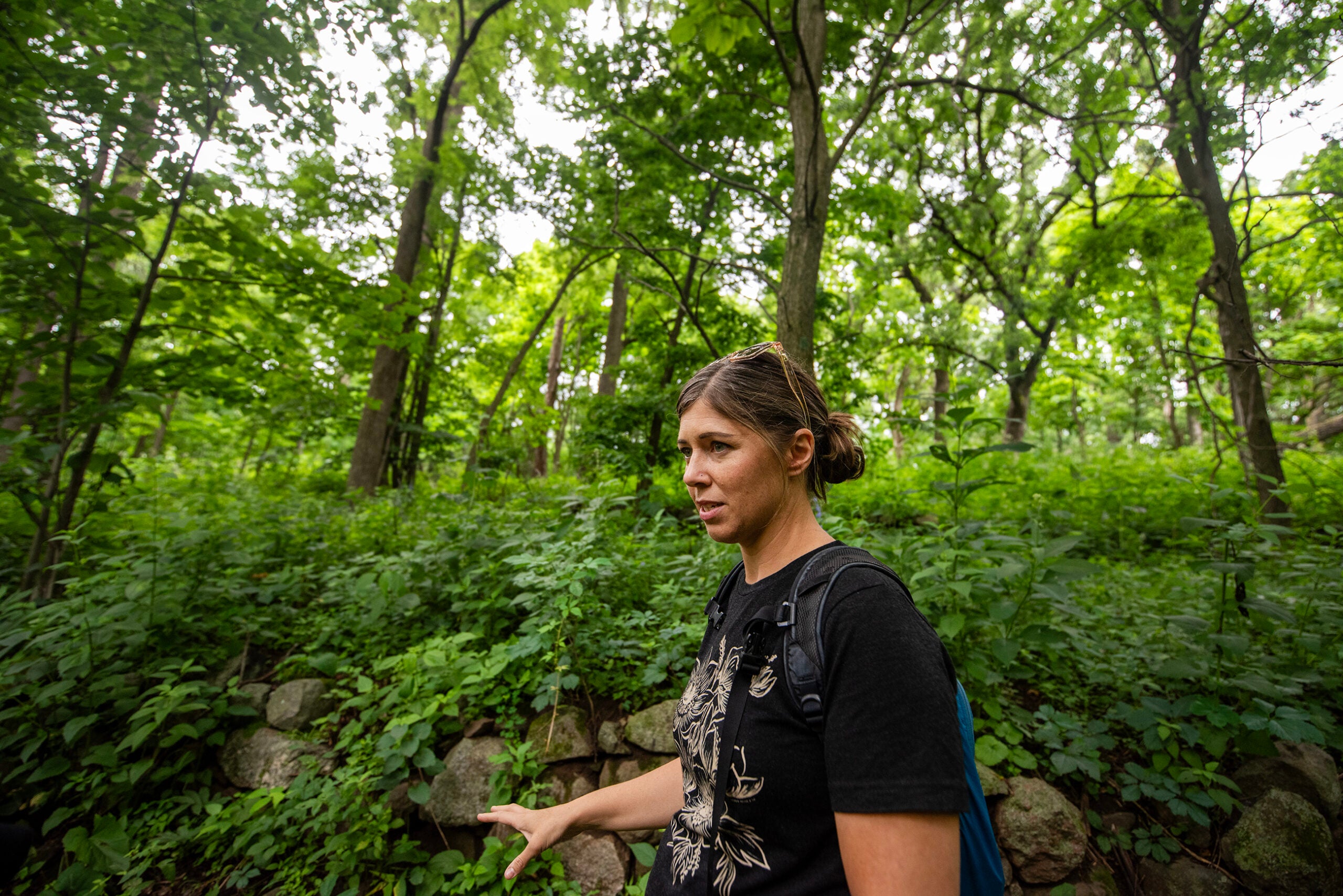 A woman with a backpack walks through a dense green forest, with a stone wall and tall trees in the background.
