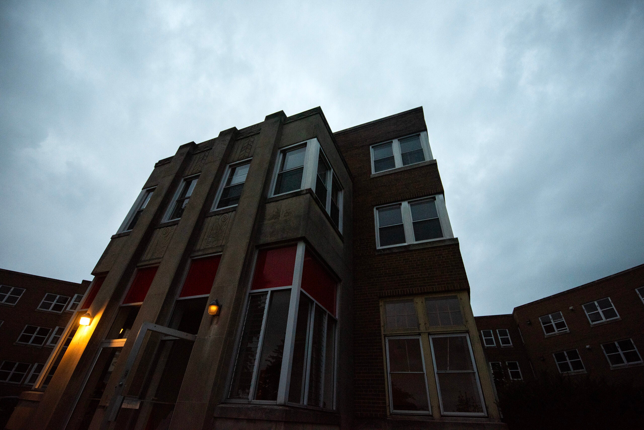 A three-story brick and concrete building with several windows, viewed from below against a cloudy sky at dusk with outdoor lights turned on.