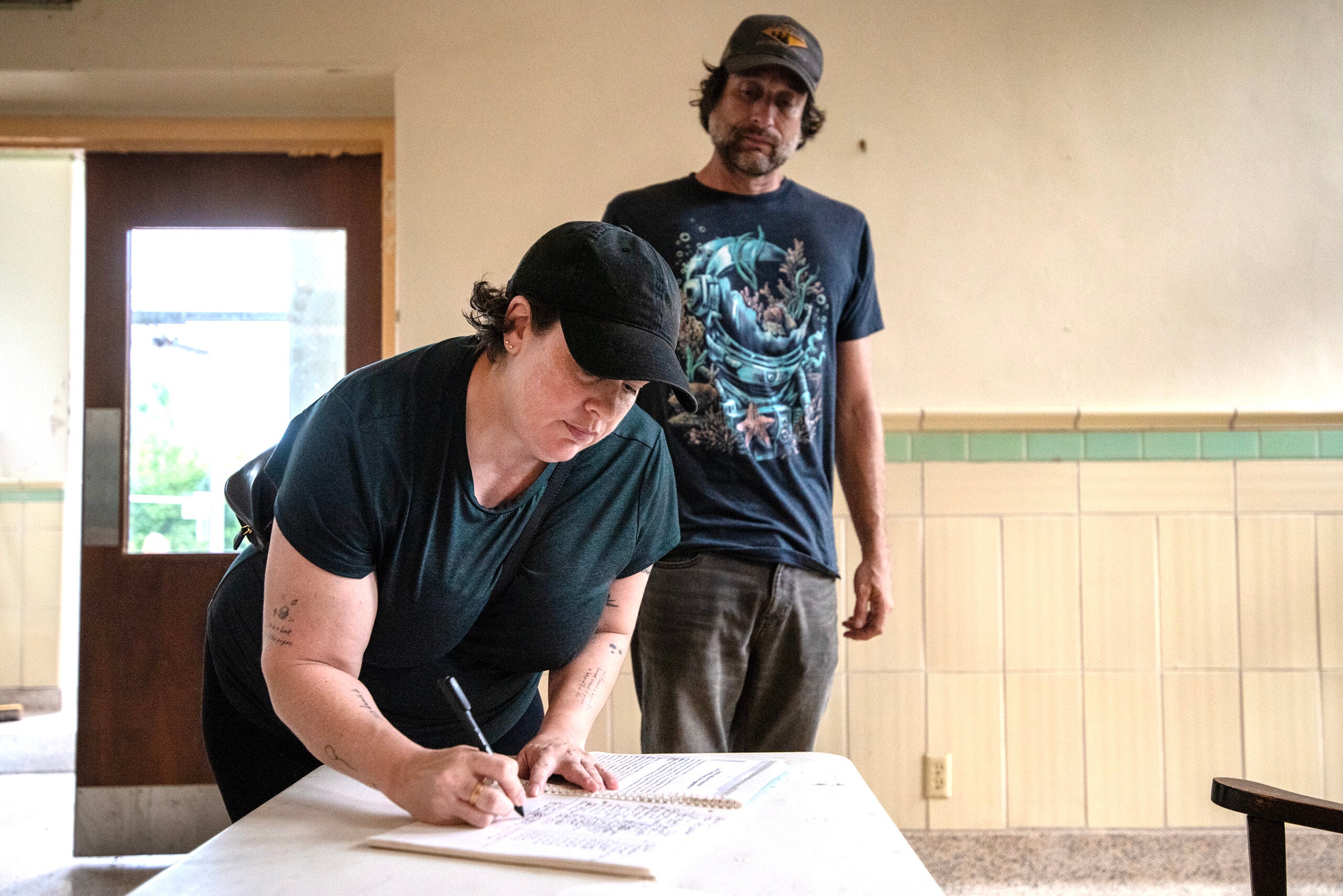 A person signs a document on a table while another person stands behind them in a room with tiled walls and a wooden door.