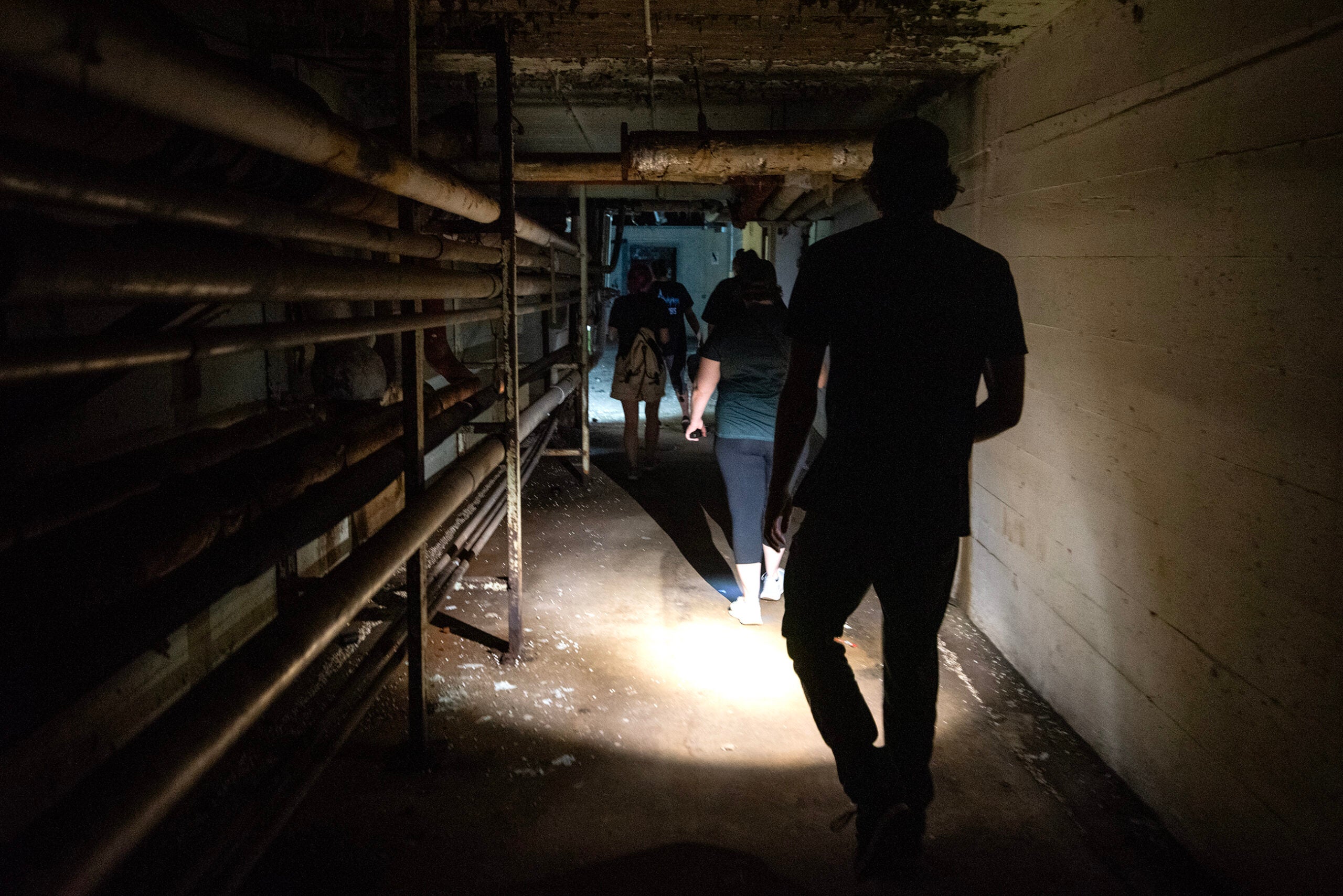 Several people walk through a dimly lit, narrow concrete hallway with exposed pipes on the left wall.