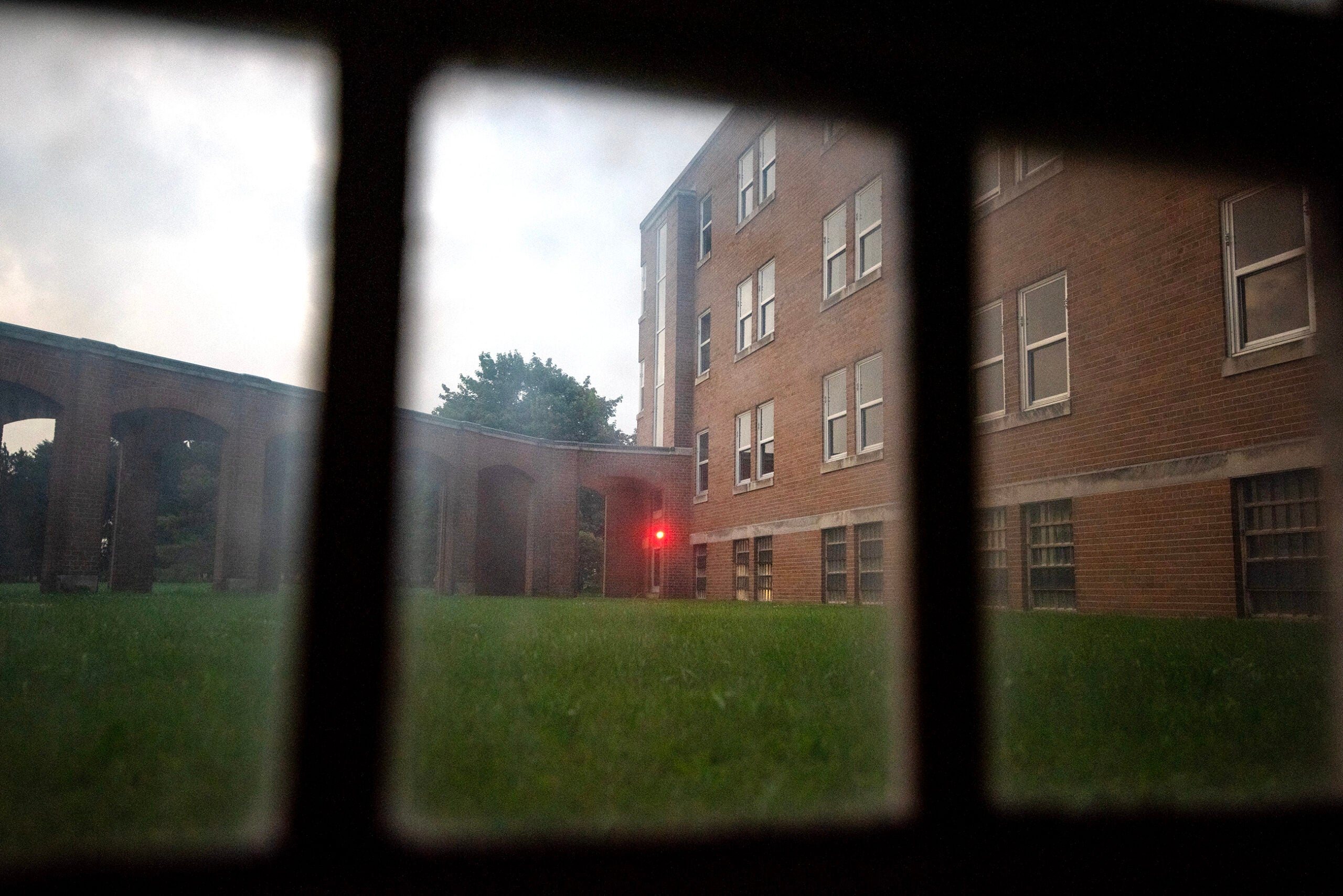 View through a window shows a grassy courtyard, brick building, and a faint red light near an exterior walkway at dusk.