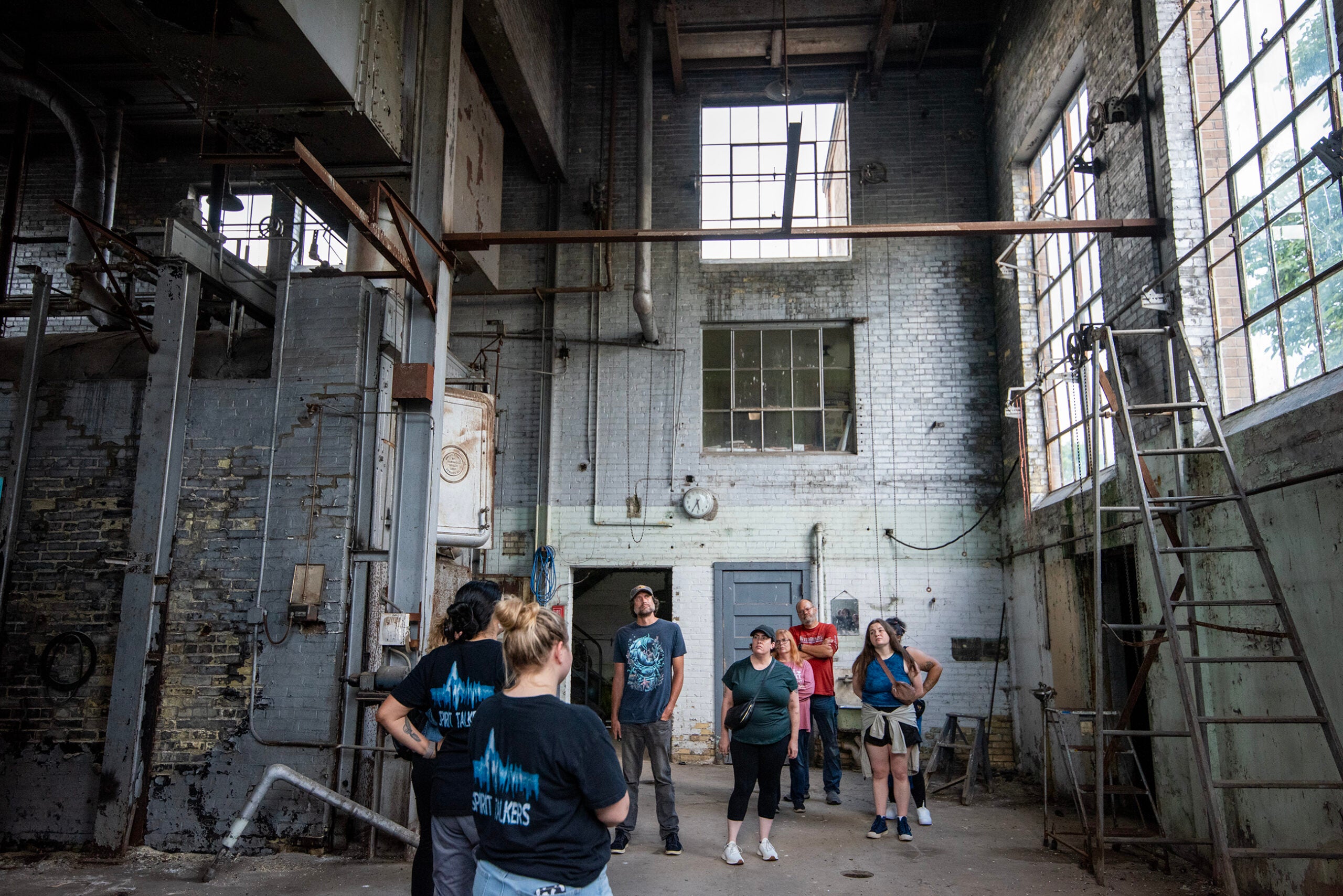 A group of people stand and talk inside an old, industrial building with worn walls, large windows, and exposed pipes.