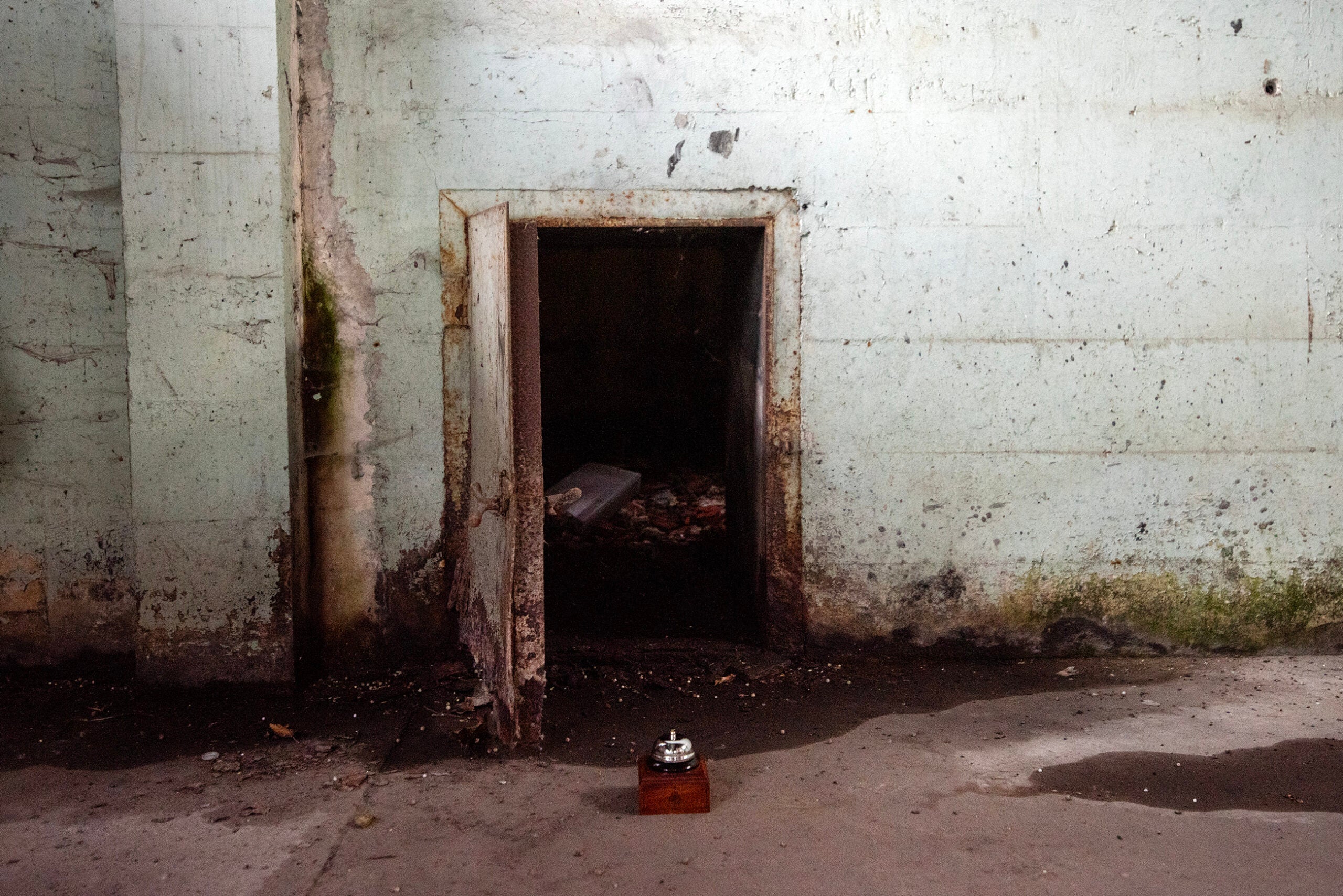 A small open doorway in a worn, dirty concrete wall leads to a dark room. A wooden box with a metal object sits on the floor in front of the door.